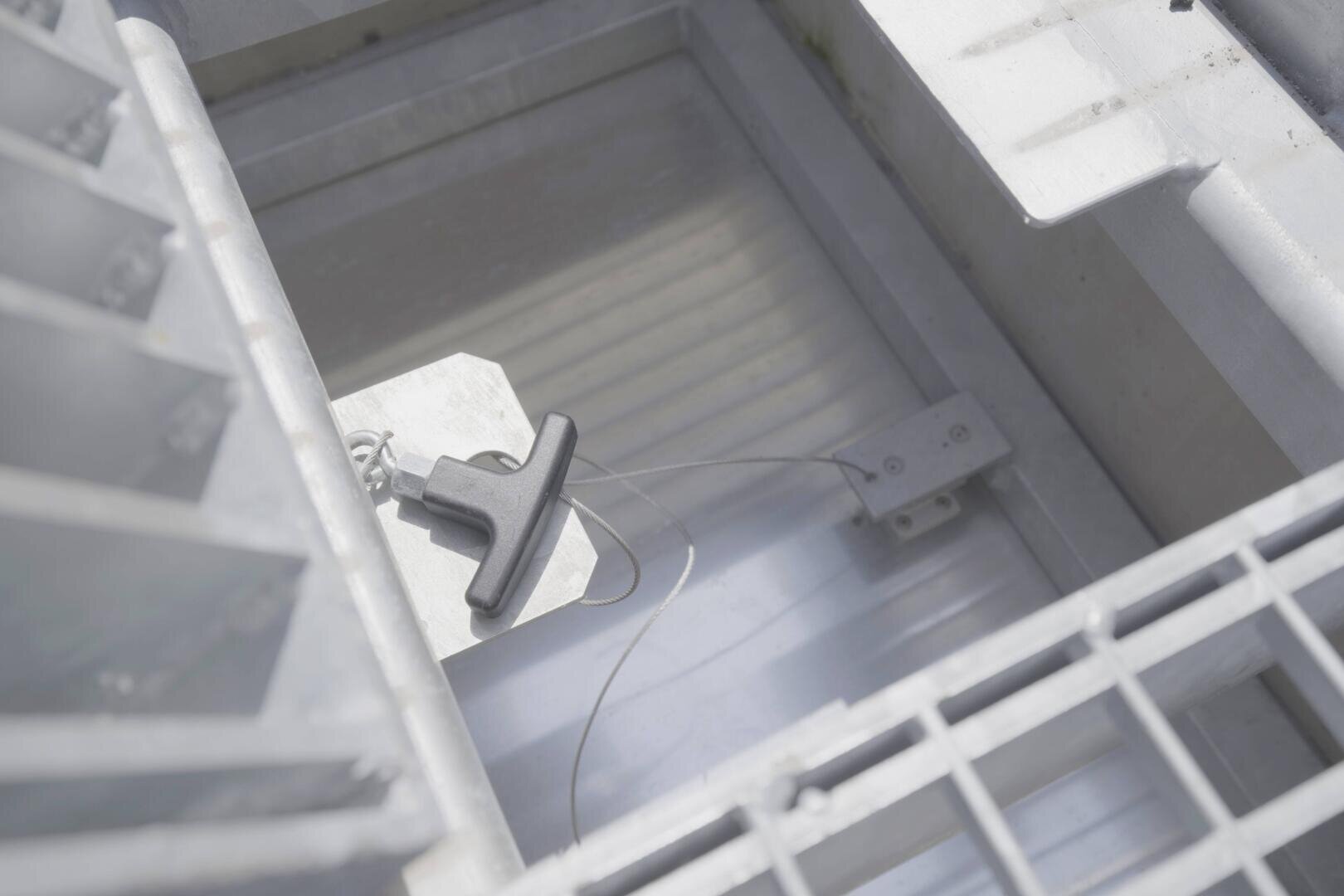 Gros plan sur une grille métallique et des récipients contenant un outil noir en forme de T, fixé à un fil fin et reposant dans un compartiment métallique rectangulaire. La scène est bien éclairée, avec des ombres et des reflets.