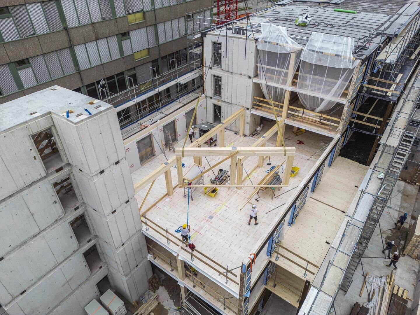 Luftaufnahme eines im Bau befindlichen mehrstöckigen Gebäudes. Zu sehen sind Sichtbetonwände, Holzbalken, Gerüste und Arbeiter auf der Baustelle. Einige Abschnitte sind mit Plastikplanen abgedeckt, und die Bauwerkzeuge liegen verstreut herum.