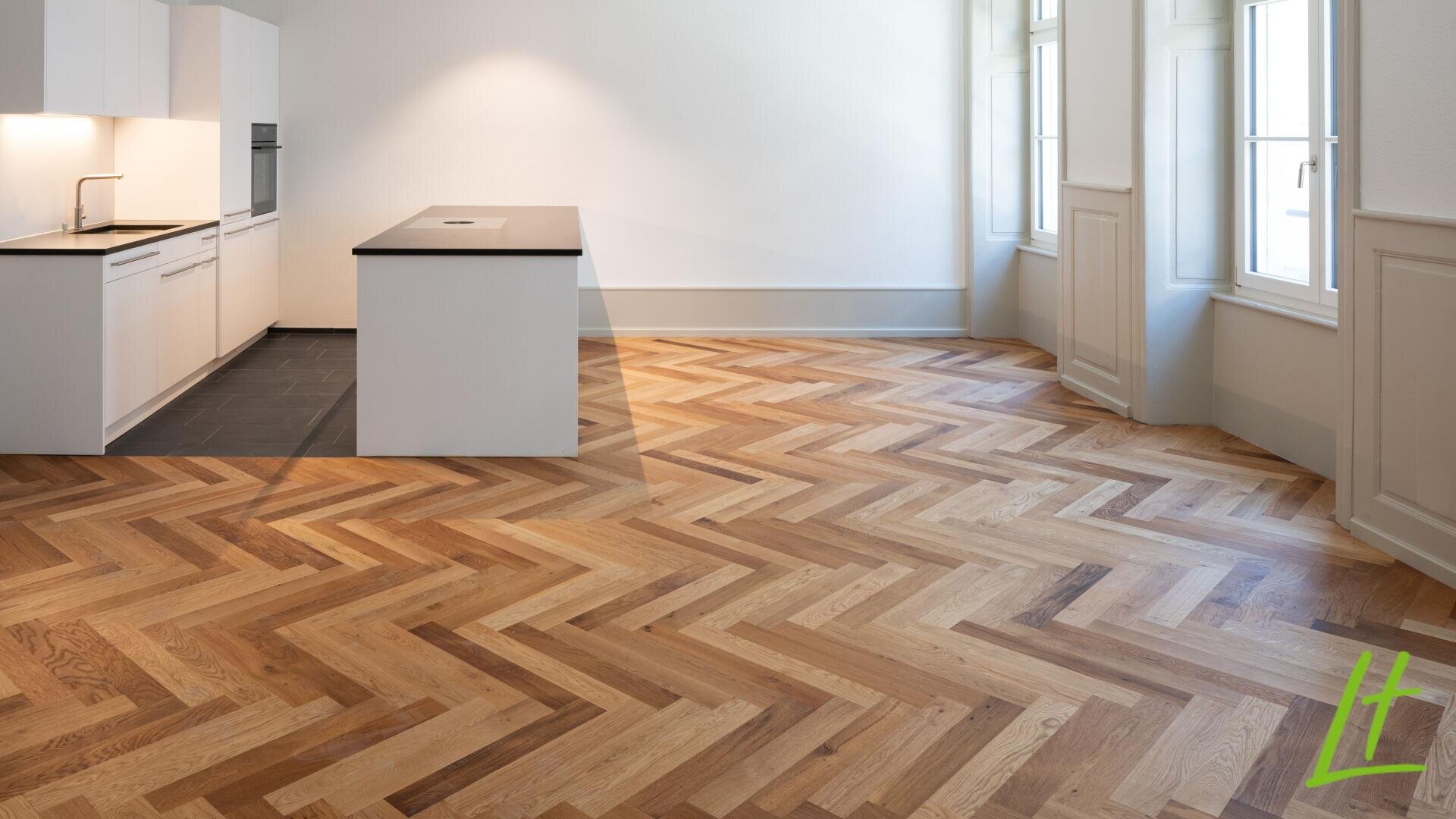 Moderne, leere Küche und Essbereich mit weißen Schränken, schwarzen Arbeitsplatten und einer zentralen Insel. Der Raum verfügt über große Fenster und helle Holzböden mit Fischgrätmuster.