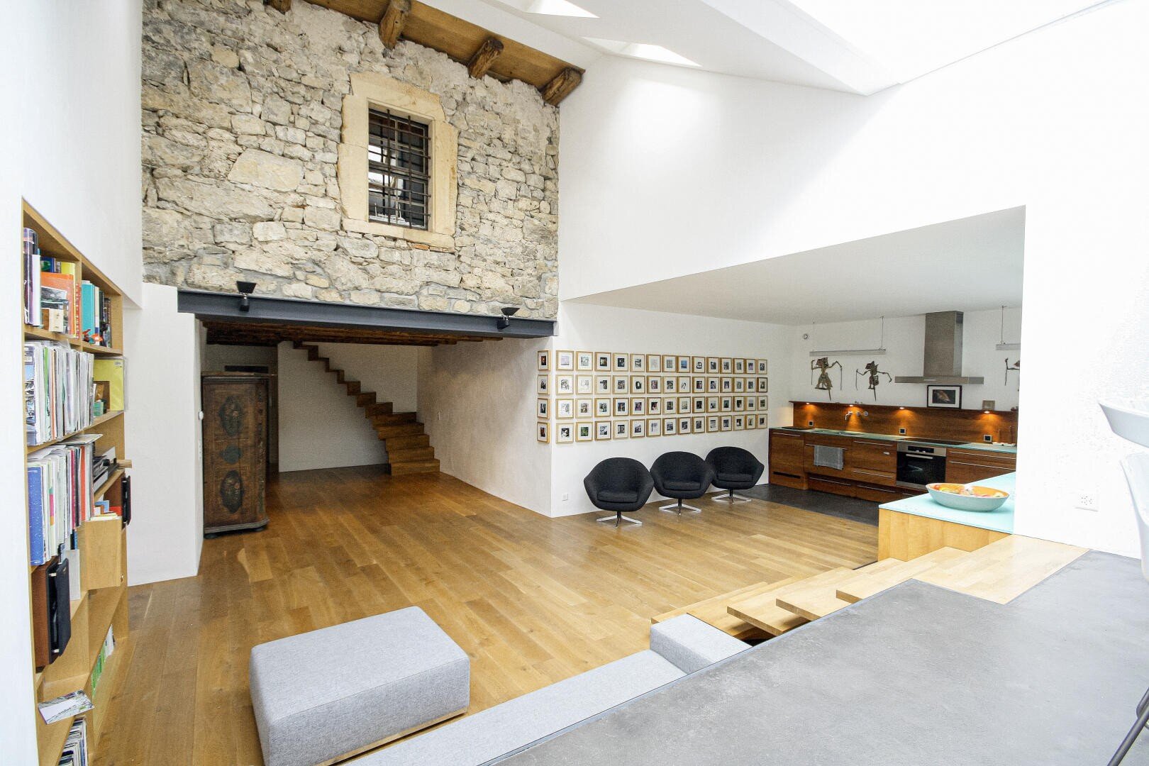 Un salon moderne ouvert avec des sols en bois, un mur en pierre exposé, une bibliothèque encastrée, quatre chaises noires, un coin cuisine et un escalier menant à un étage supérieur avec une fenêtre rustique.