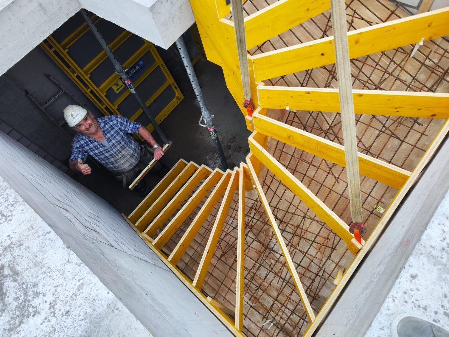 Ein Bauarbeiter mit Schutzhelm zeigt den Daumen nach oben neben einer teilweise gebauten Betontreppe mit gelber Holzschalung und freiliegender Bewehrungsstruktur.
