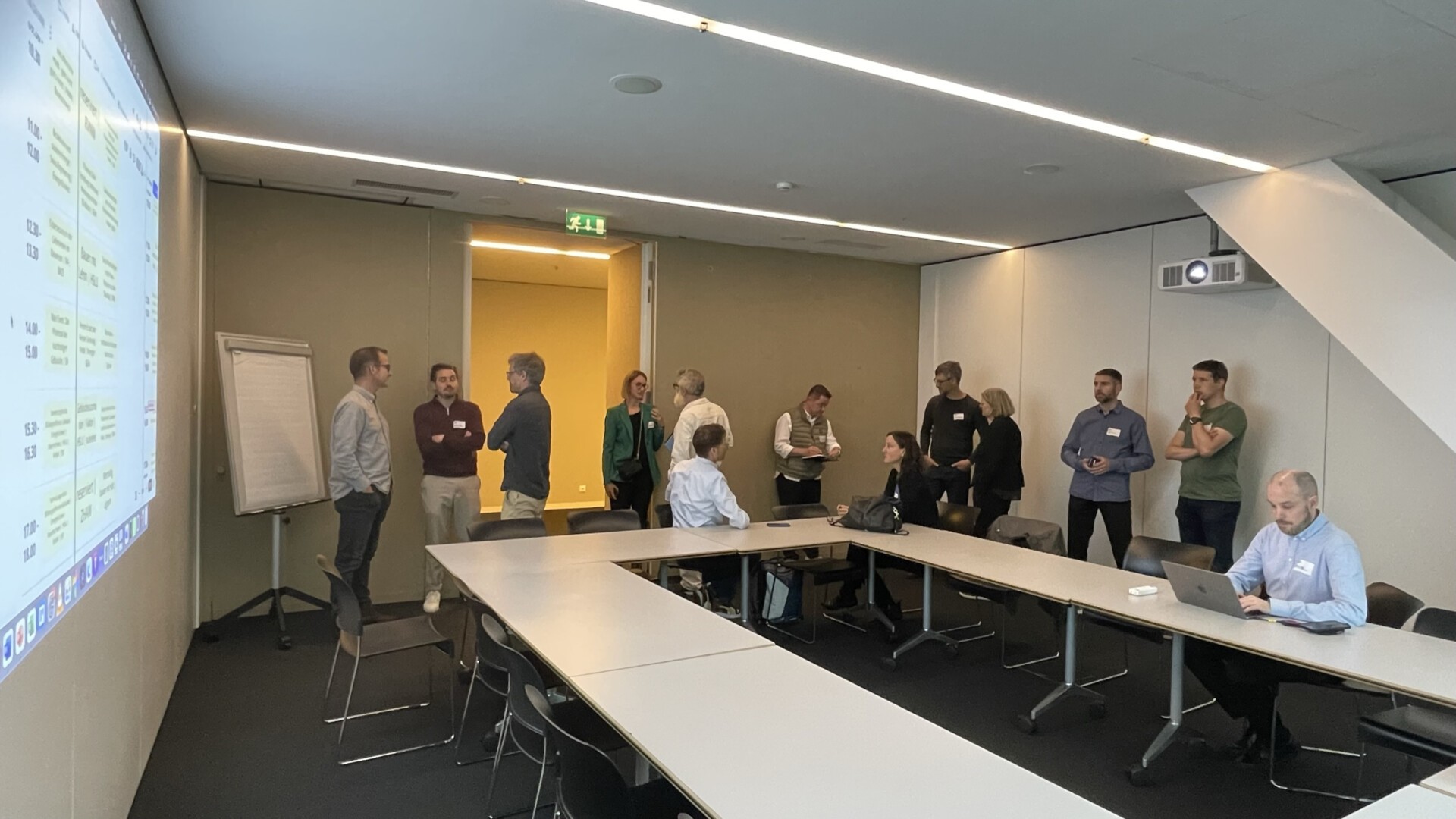 Eine Gruppe von Menschen steht und unterhält sich in einem Konferenzraum neben einer Projektionsleinwand, auf der ein Zeitplan angezeigt wird, während einige Personen mit Laptops und Notizblöcken an Tischen sitzen.