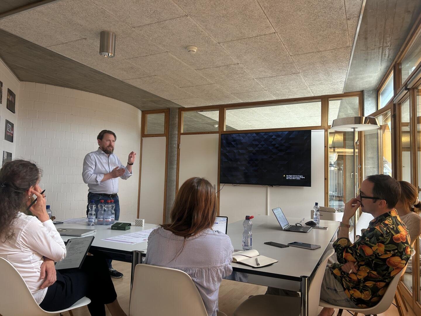 Ein Mann steht und präsentiert vier Personen, die um einen Konferenztisch mit Laptops und Papieren sitzen; hinter ihm ist ein großer Bildschirm an der Wand in einem modernen Besprechungsraum mit Tageslicht angebracht.