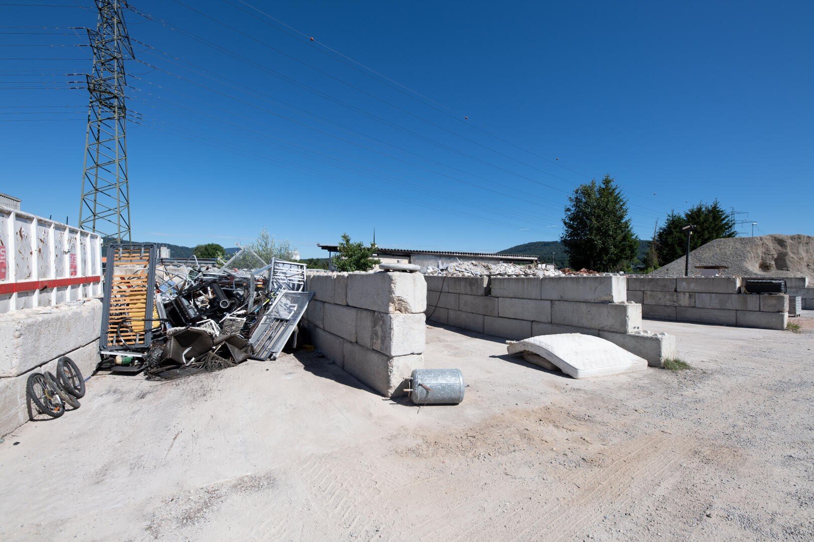 Ein Bau- oder Industriegelände mit verstreuten Trümmern, Betonblöcken, Metallresten und Holzpaletten unter einem klaren blauen Himmel. Im Hintergrund sind Stromleitungen und Bäume zu sehen.