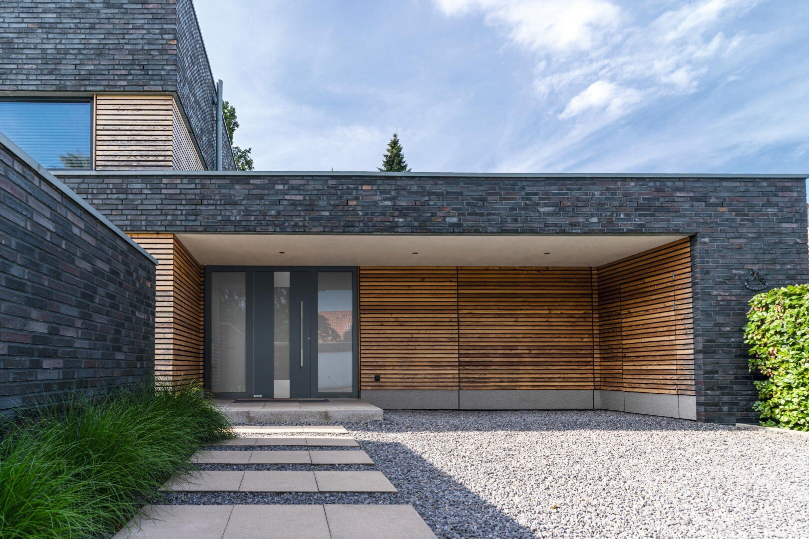Ein moderner Hauseingang mit dunklem Ziegelstein und horizontalen Holzlatten, großen Doppeltüren mit Glasscheiben, Kiesauffahrt, Steinweg und grünen Sträuchern an der Seite.