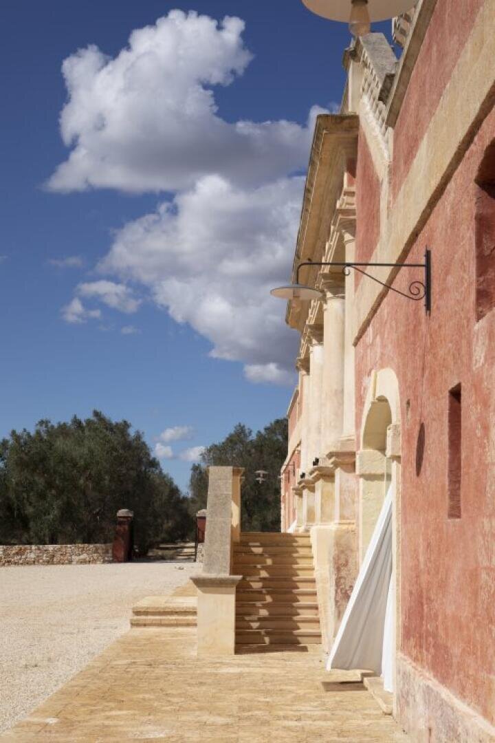 Un bâtiment rustique en pierre avec des murs rouges et beiges, de larges marches et des détails en fer forgé se détache sur un ciel bleu vif avec des nuages blancs et floconneux. Des oliviers et un chemin de gravier sont visibles à l'arrière-plan.