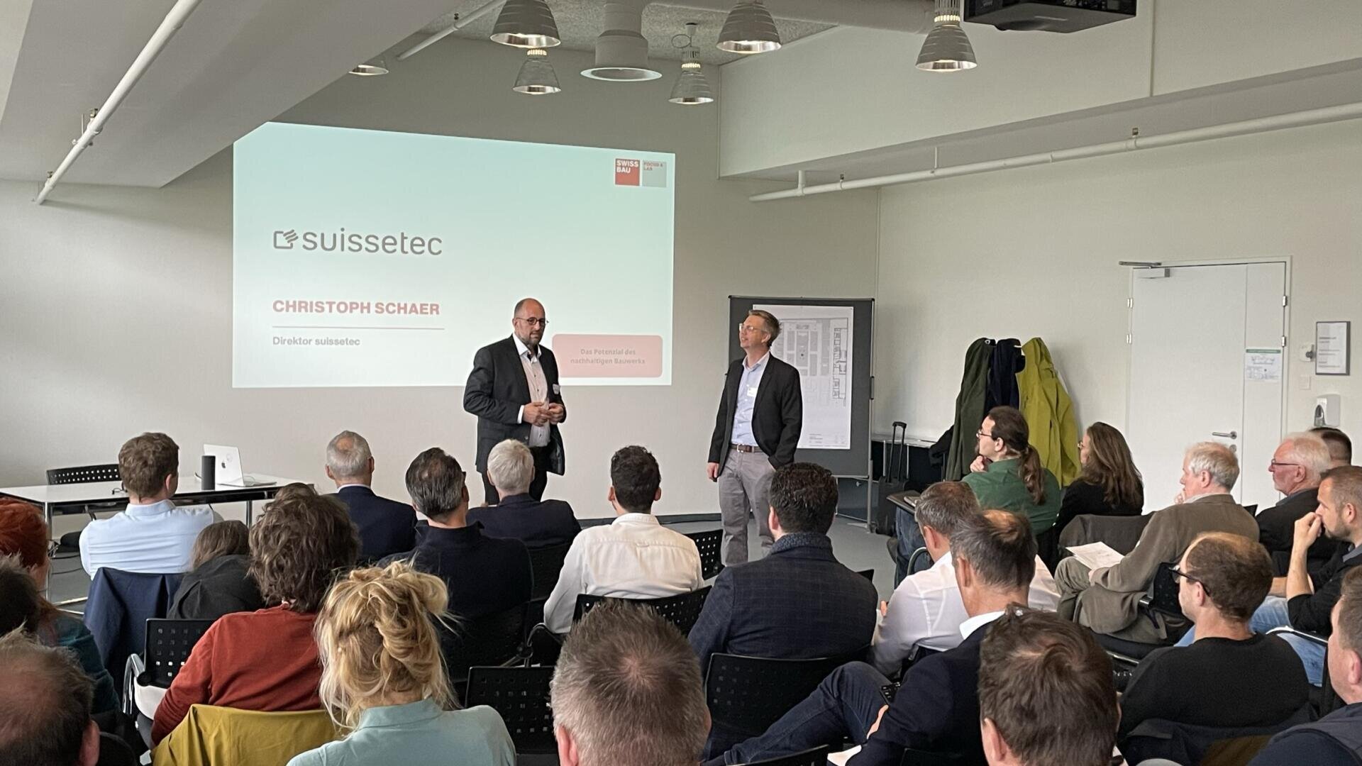 Ein Mann im Anzug spricht in einem Konferenzraum zu seinem Publikum. Hinter ihm ist eine Präsentationsfolie zu sehen, auf der suissetec und Christoph Schaer zu sehen sind. Ein weiterer Mann steht neben ihm, und die Anwesenden hören aufmerksam zu.