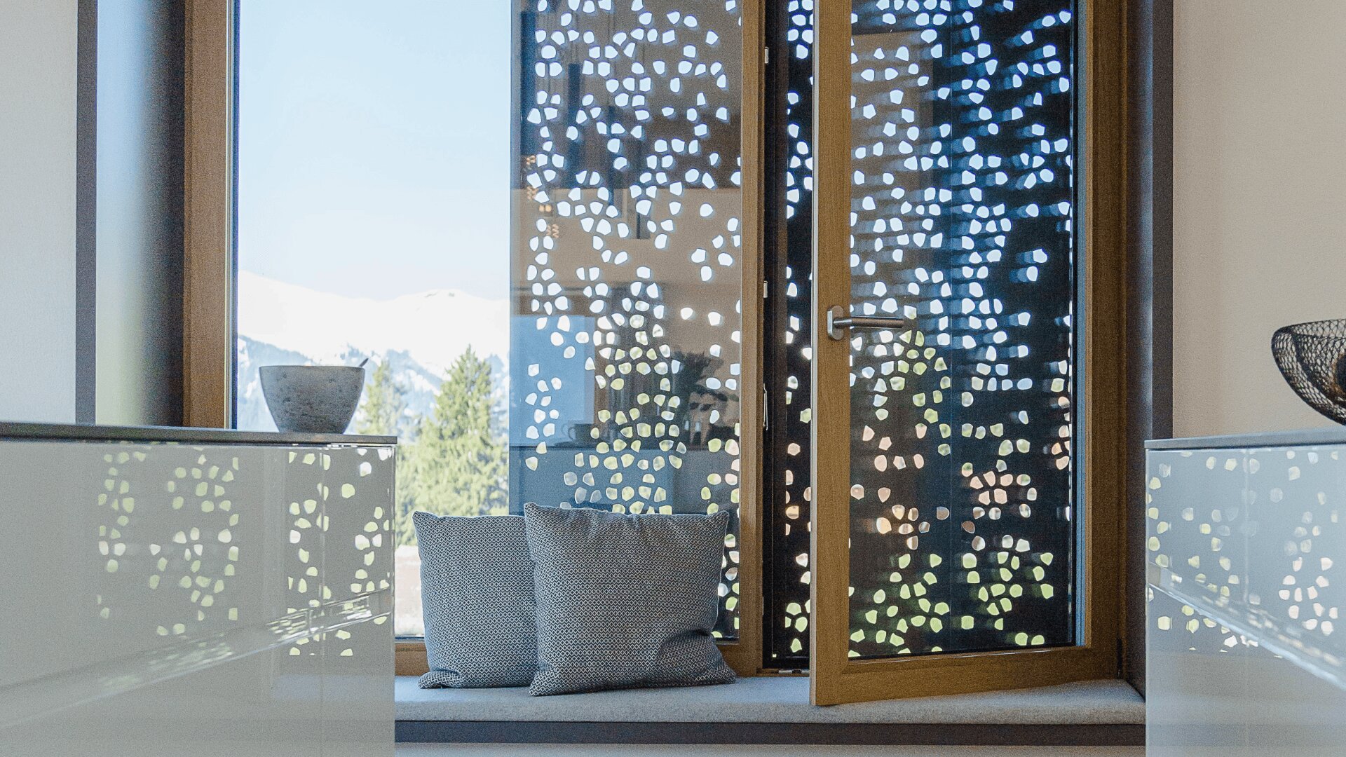 Ein Fenster mit einem gemusterten Sichtschutz lässt das Sonnenlicht und die Außenlandschaft durch. Zwei Kissen liegen auf der Fensterbank, und das moderne Dekor spiegelt sich in den glänzenden Oberflächen auf beiden Seiten wider.