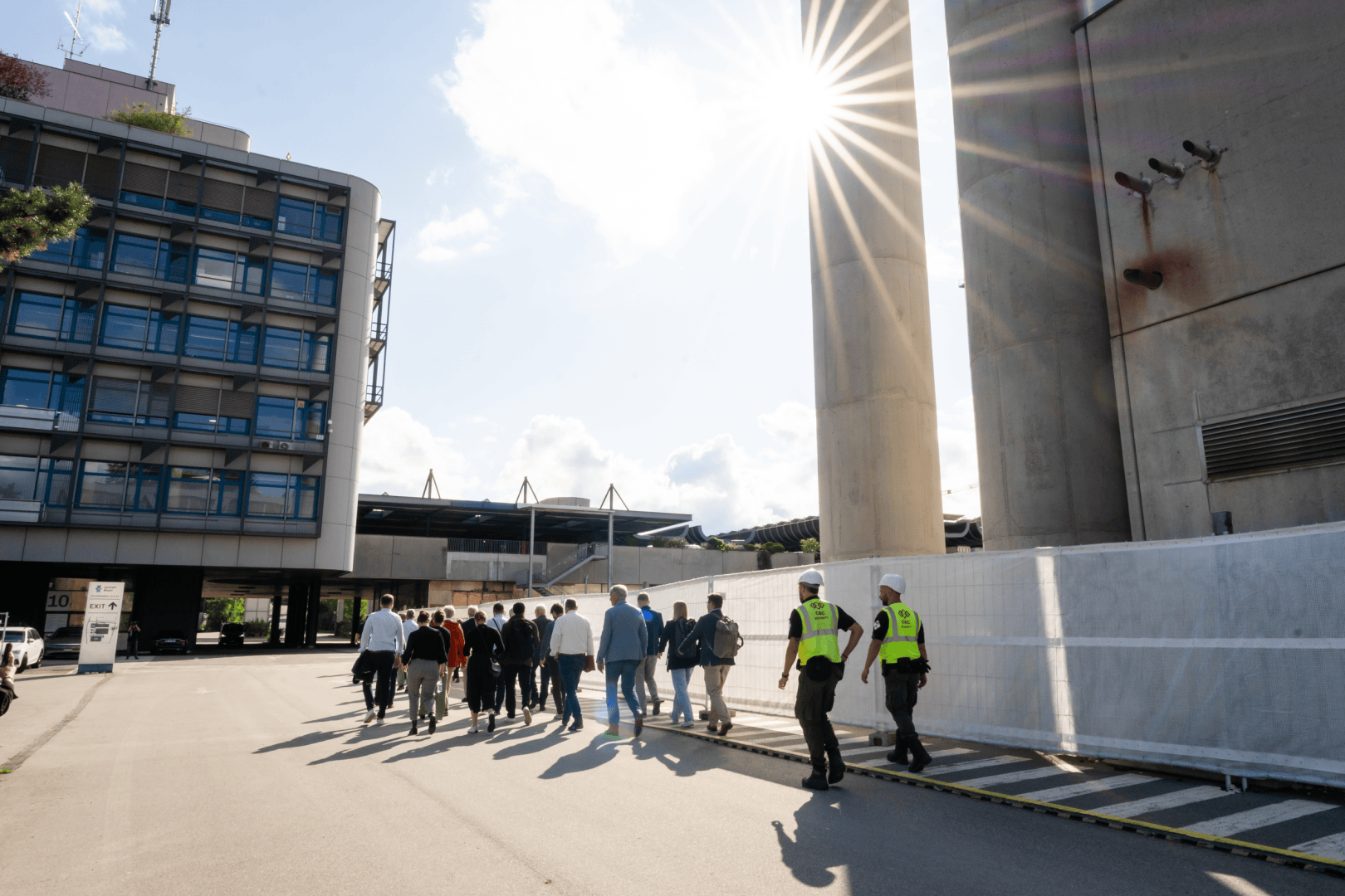 Eine Gruppe von Menschen geht gemeinsam auf einer Straße zwischen modernen Gebäuden bei strahlendem Sonnenschein, während zwei Sicherheitsbeamte in der Nähe beobachten.