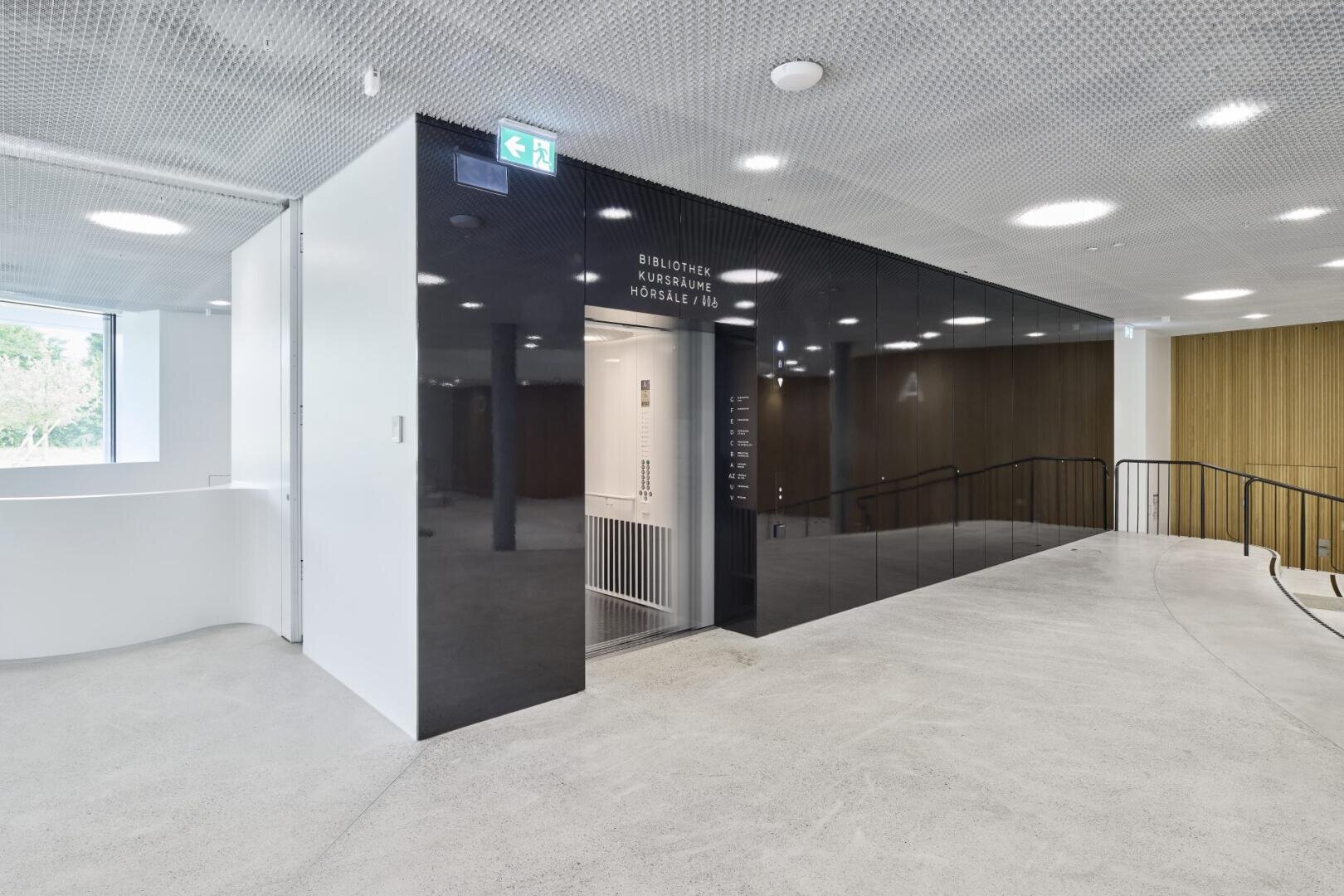 Modernes, minimalistisches Interieur mit schwarzem Aufzug, weißen Wänden und hellgrauem Bodenbelag. Ein Schild über dem Aufzug listet die Standorte auf, und daneben befindet sich eine gebogene Rampe. Große Fenster lassen viel natürliches Licht herein.