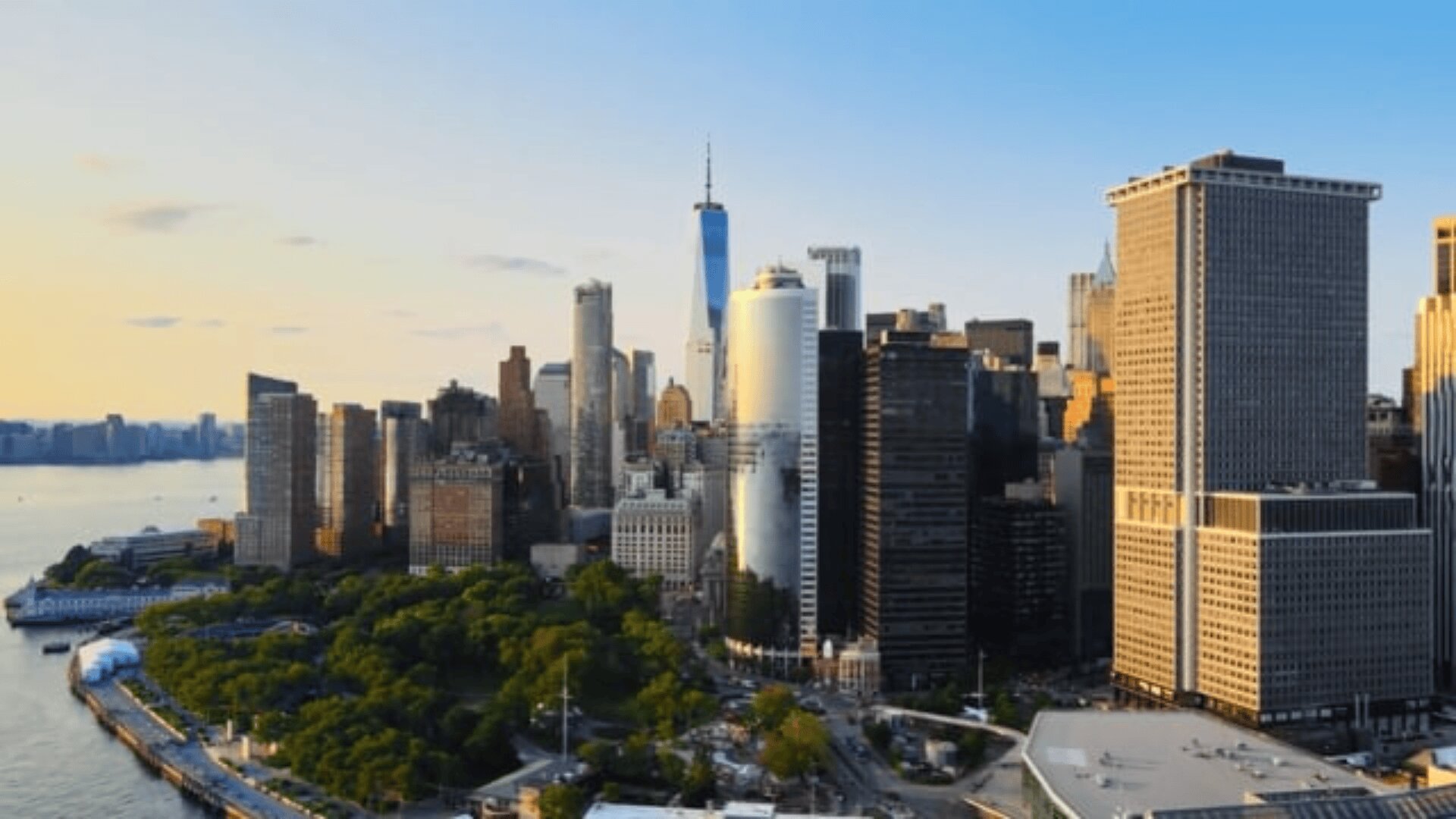 Luftaufnahme von Lower Manhattan in New York City bei Sonnenuntergang, mit hohen Wolkenkratzern, dem One World Trade Center und einem Park am Wasser mit Bäumen im Vordergrund.