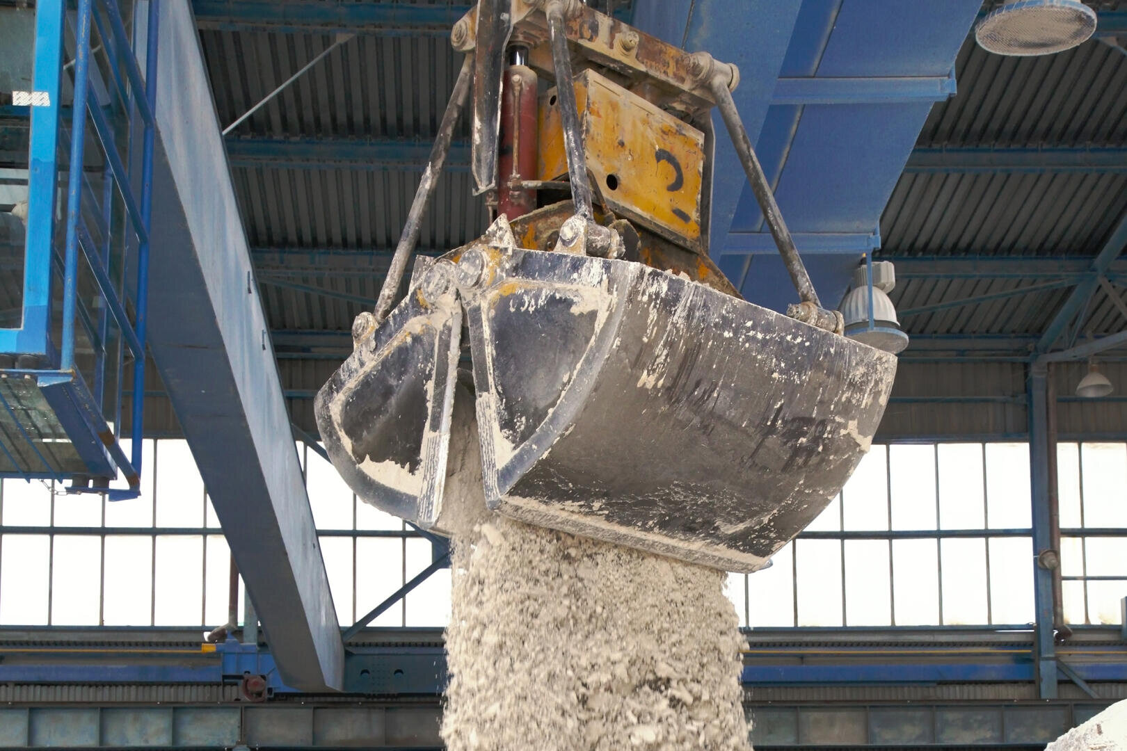 Ein großer industrieller Zweischalengreifer gibt eine Ladung Kies oder Sand in einer Fabrik oder einem Lagerhaus mit blauen Stahlträgern und hohen Fenstern im Hintergrund frei.