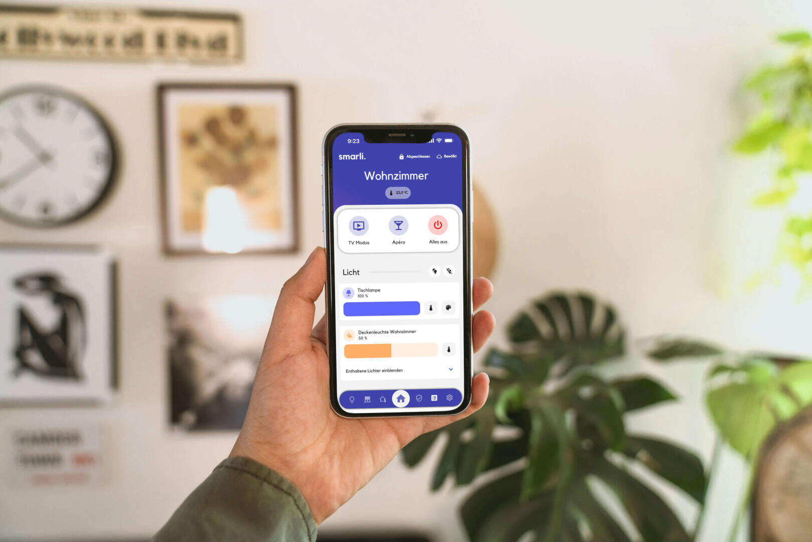 Eine Person hält ein Smartphone in der Hand, auf dem eine deutschsprachige Smart-Home-App angezeigt wird, die Steuerungen für TV/HiFi, Alarm und Beleuchtung im Wohnzimmer anzeigt. Im Hintergrund sind Zimmerpflanzen, Wandkunst und eine Uhr zu sehen.