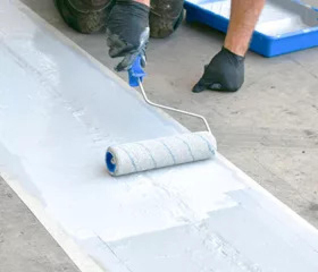 Eine Person mit schwarzen Handschuhen trägt mit einem Farbroller weiße Farbe auf einen Bodenstreifen auf, wobei im Hintergrund eine Farbwanne zu sehen ist.