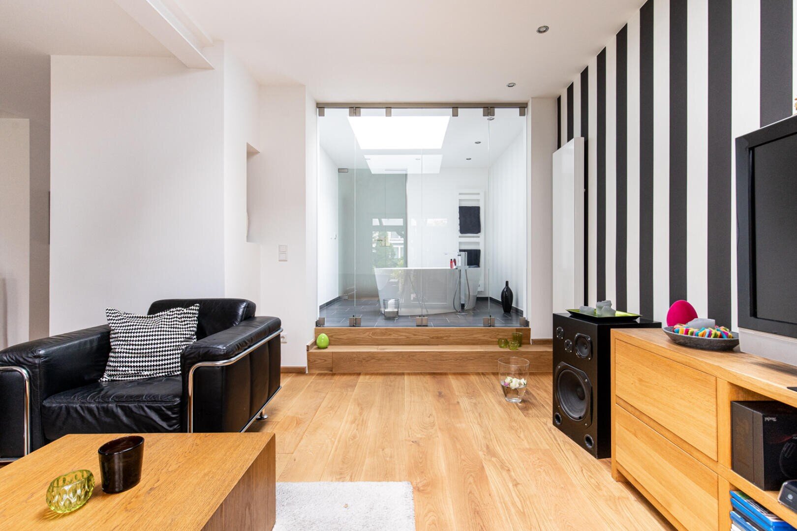 Modernes Wohnzimmer mit Holzböden, einem schwarzen Ledersessel, einer gestreiften Akzentwand und einem Blick durch Glastüren in eine helle, weiße Küchenzeile. Minimalistisches Dekor und zeitgenössische Möbel sind hier zu finden.