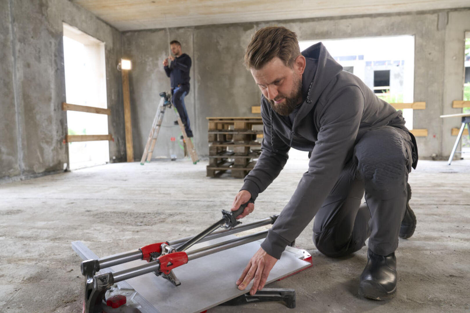 Un homme agenouillé sur le sol découpe un carreau de céramique à l'aide d'un coupe-carreaux dans une pièce inachevée, tandis qu'un autre homme, debout sur une échelle, travaille en arrière-plan.