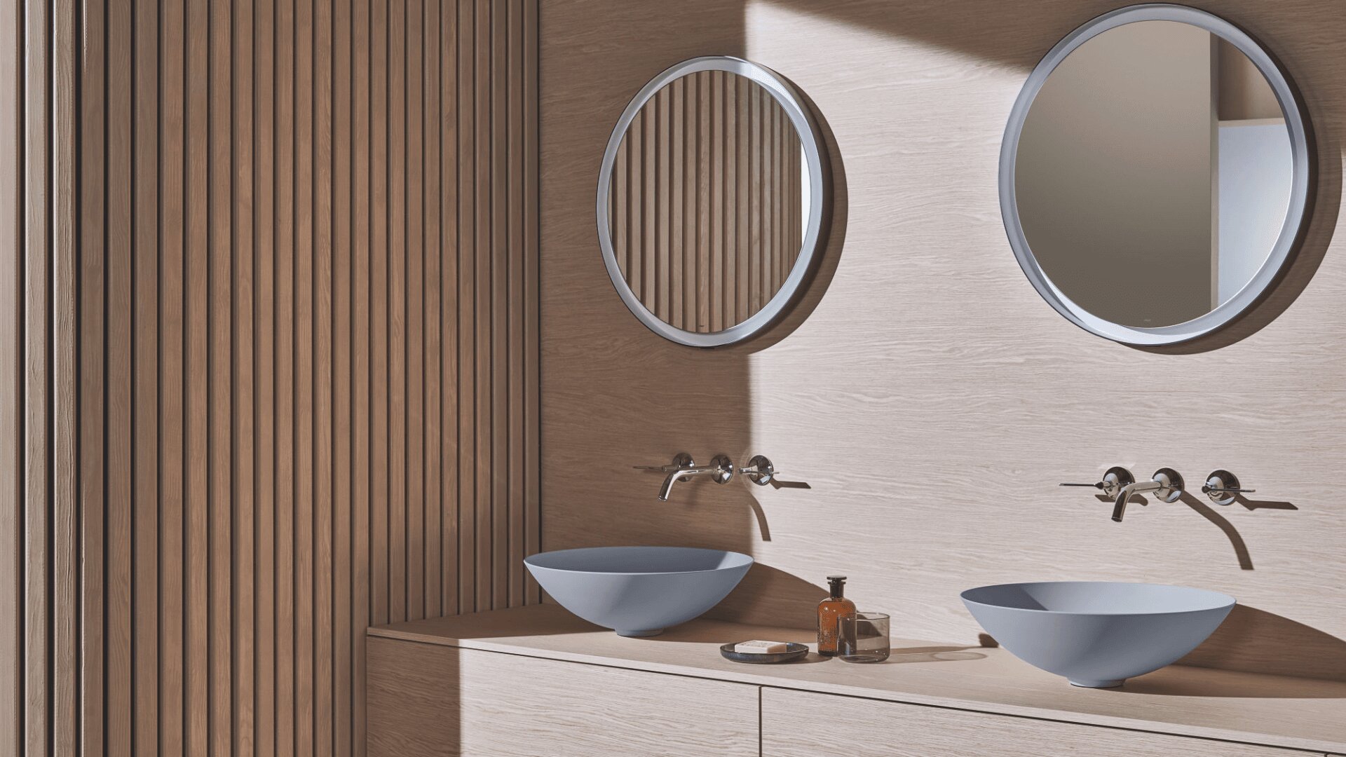 Ein modernes Badezimmer mit hellen Holzpaneelen, zwei runden Spiegeln, zwei blauen Waschbecken, Wandarmaturen, Seife und einer Flasche auf der Theke. Das Sonnenlicht wirft diagonale Schatten auf die Oberflächen.