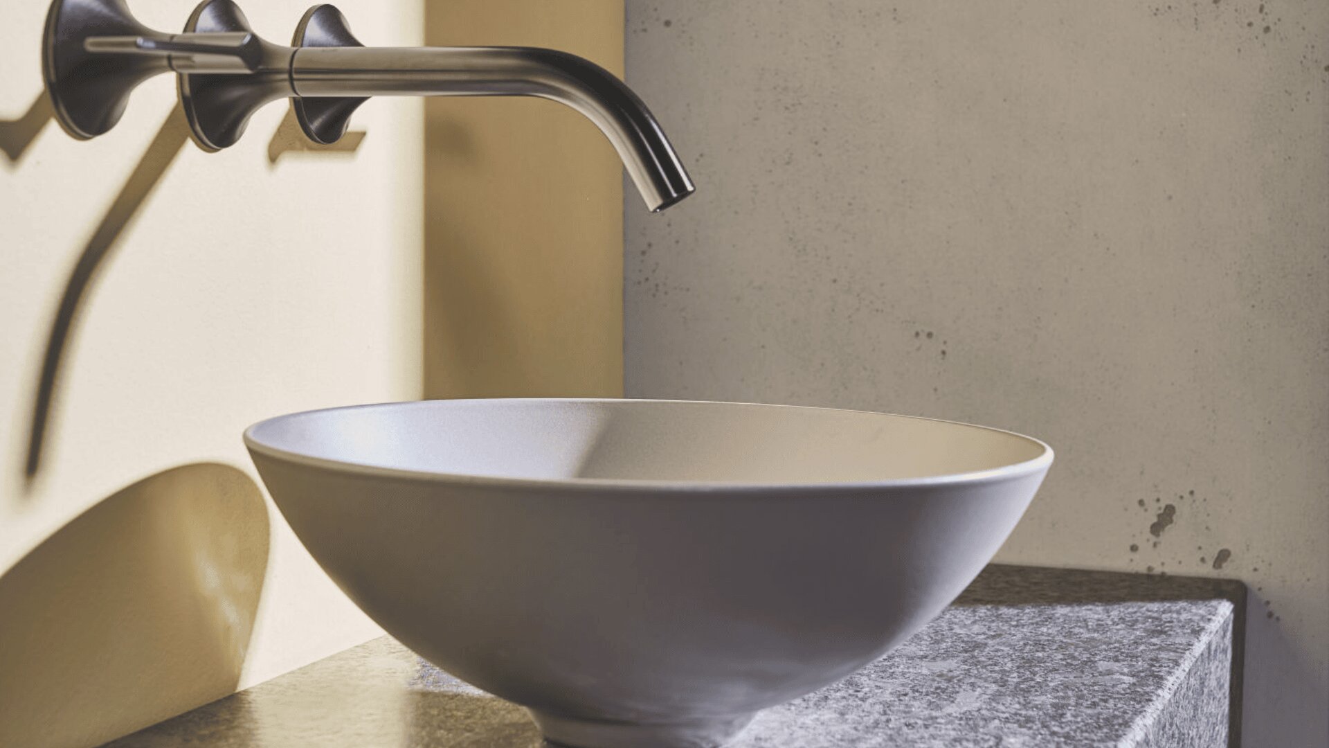 In einem modernen Badezimmer steht eine runde weiße Spüle auf einer Granitarbeitsplatte mit einem eleganten Metallwasserhahn an der Wand neben einer hellfarbigen, strukturierten Wand.