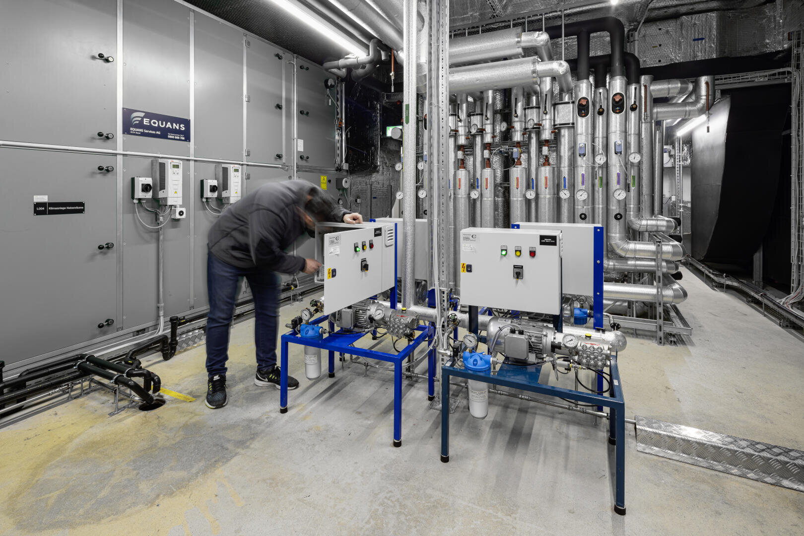 Eine Person inspiziert Schalttafeln in einer modernen Industrieanlage mit freiliegenden Rohren, Schaltkästen und Maschinen. Der Raum ist gut beleuchtet und geordnet, mit Etiketten und Sicherheitsschildern versehen.
