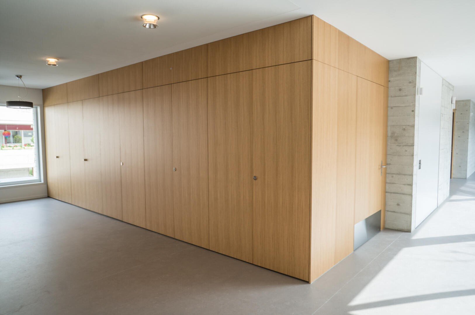 Ein moderner Innenraum mit einer großen Holzstruktur mit glatten Paneelen, eingebauten Türen und einer kleinen rechteckigen Öffnung am Boden. Der Raum hat Betonwände, große Fenster und helles Tageslicht.