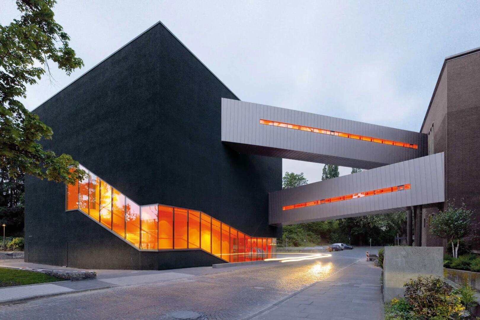 Ein modernes schwarzes Gebäude mit großen orangefarbenen Fenstern und zwei geschlossenen Brücken, die mit einem anderen Gebäude verbunden sind, beleuchtet in der Abenddämmerung, mit Bäumen und einer Kopfsteinpflasterstraße im Vordergrund.
