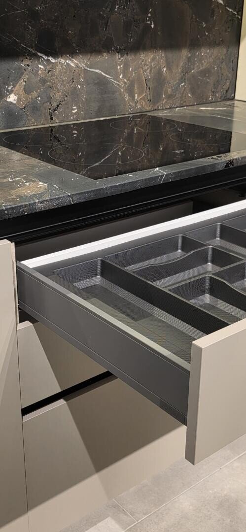 Un tiroir de cuisine ouvert avec un range-ustensiles vide, sous un comptoir en marbre noir et une cuisinière électrique. Le tiroir et les armoires sont de couleur beige clair.