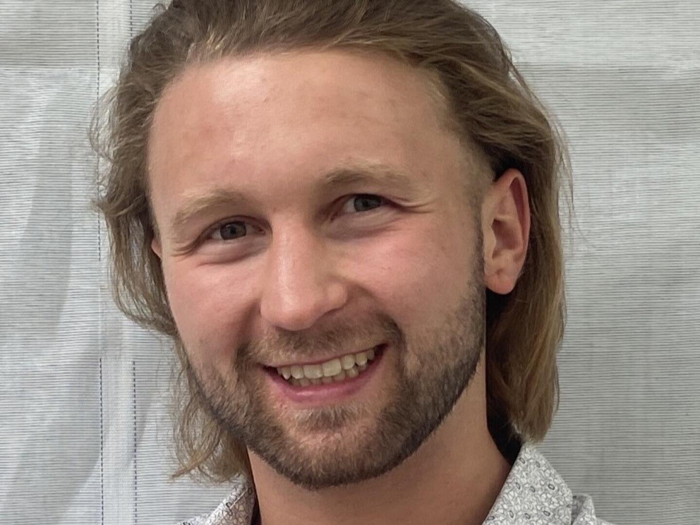 Eine Person mit langen hellbraunen Haaren, einem Bart und Schnurrbart lächelt in die Kamera. Sie trägt ein helles, gemustertes Hemd und steht vor einem hellen, strukturierten Hintergrund.