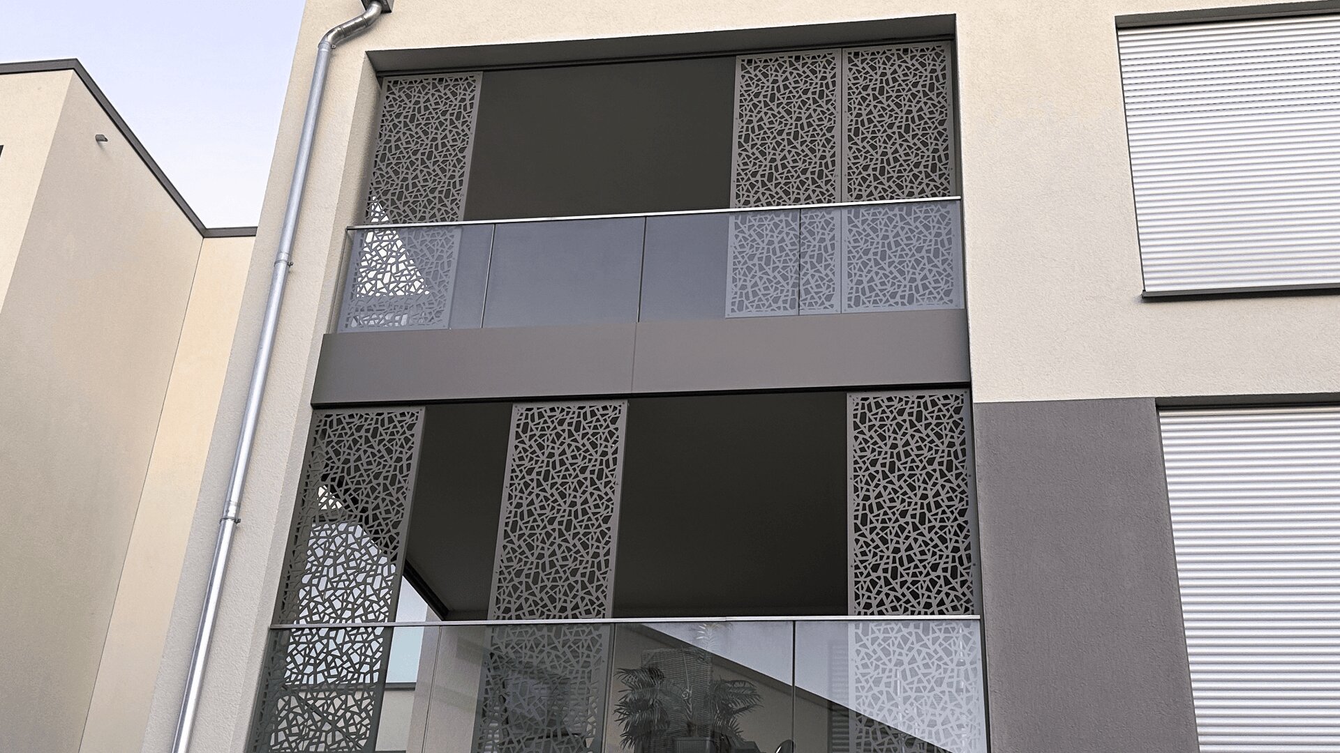 Fassade eines modernen Gebäudes mit dekorativen Metallpaneelen mit abstraktem geometrischem Muster, Glasgeländern und Rollläden an den Fenstern. Eine Topfpflanze ist durch das Glas auf einem Treppenabsatz zu sehen.