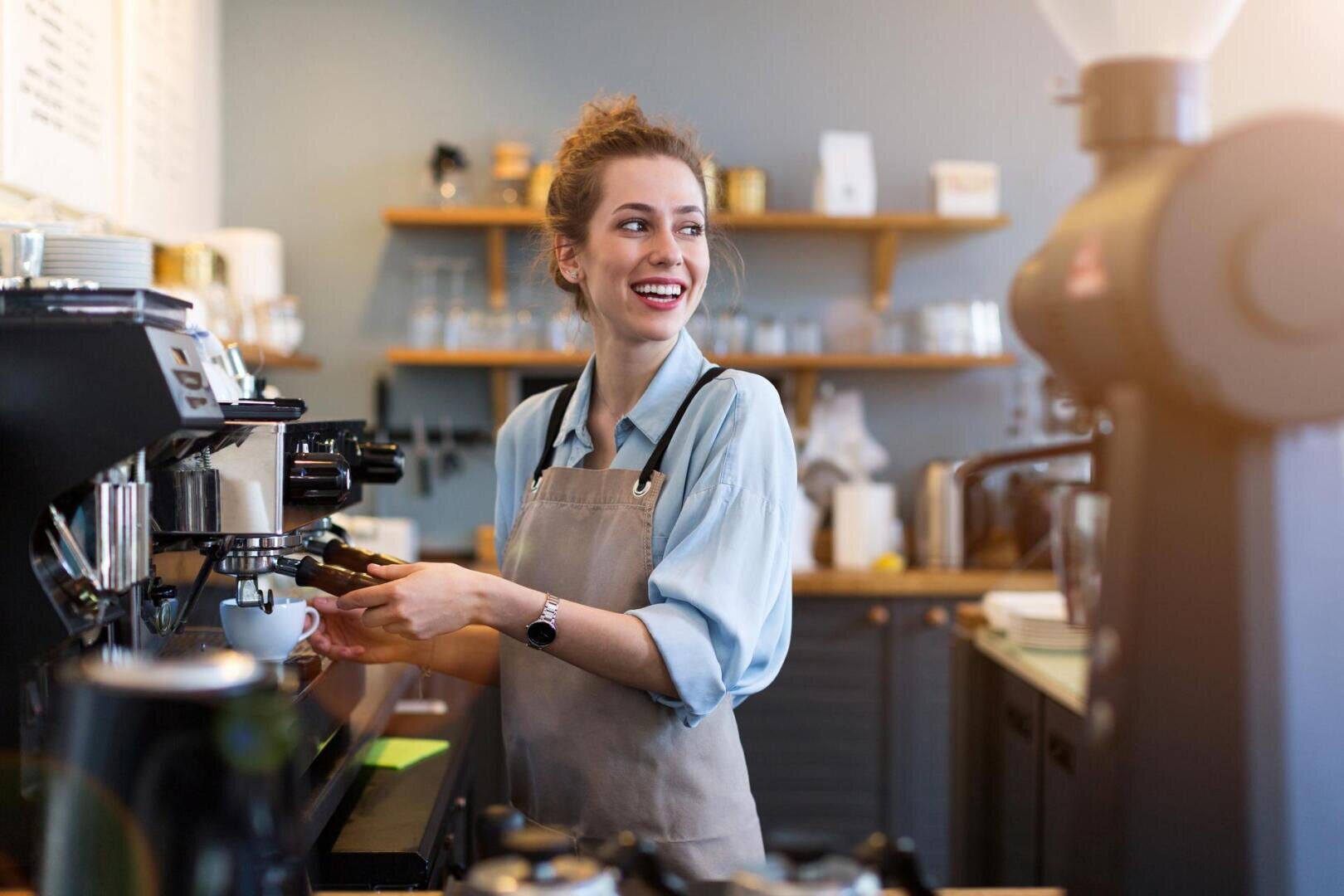Un barista souriant en chemise et tablier bleus prépare une boisson au café sur une machine à expresso dans un café moderne, avec des étagères et des tasses en arrière-plan.