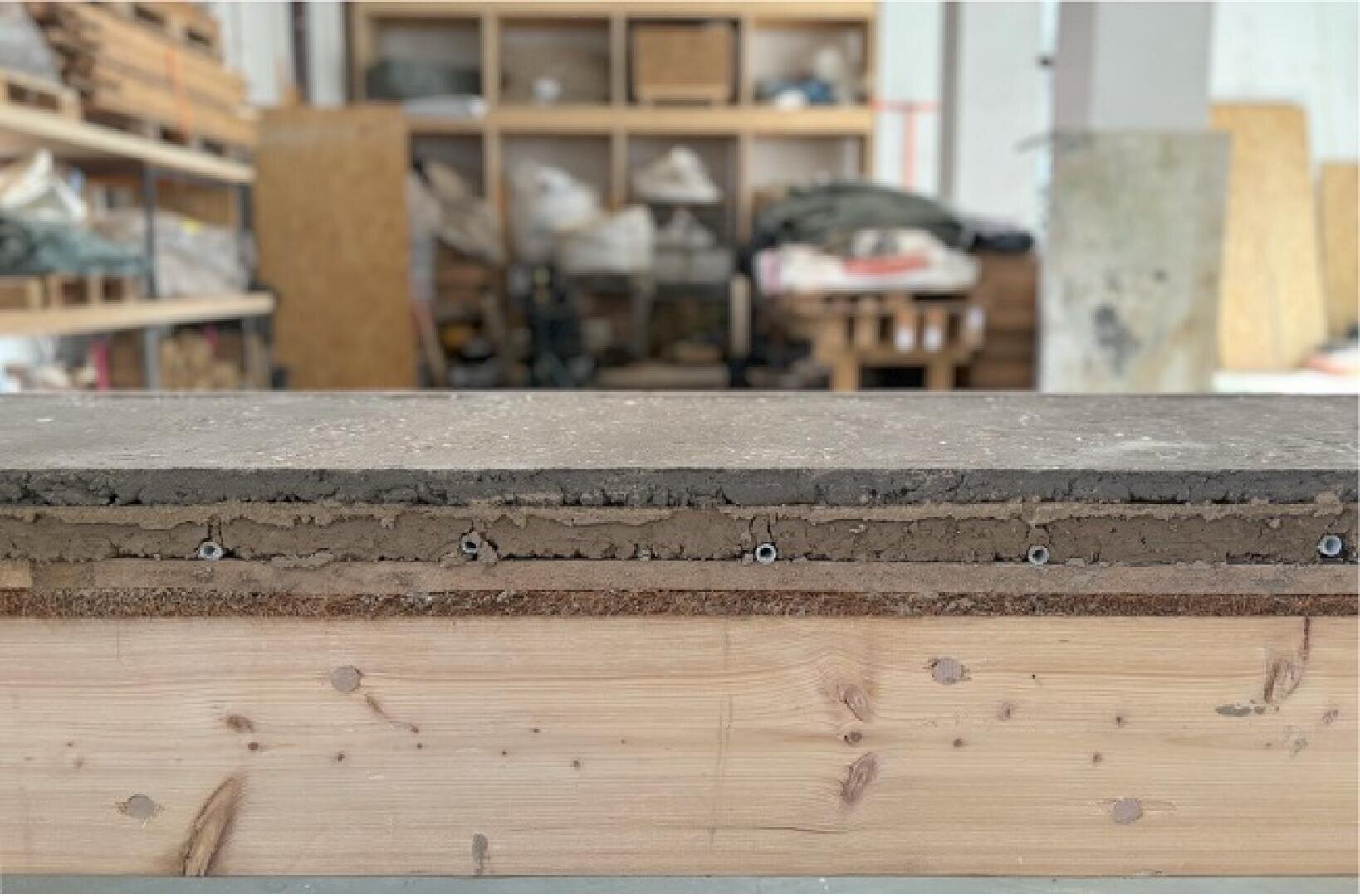 Nahaufnahme eines Querschnitts eines geschichteten Baumaterials, möglicherweise Dämmstoff oder Lehm, auf einer hölzernen Unterlage, mit Regalen und Werkzeugen, die im Hintergrund einer Werkstatt verschwimmen.
