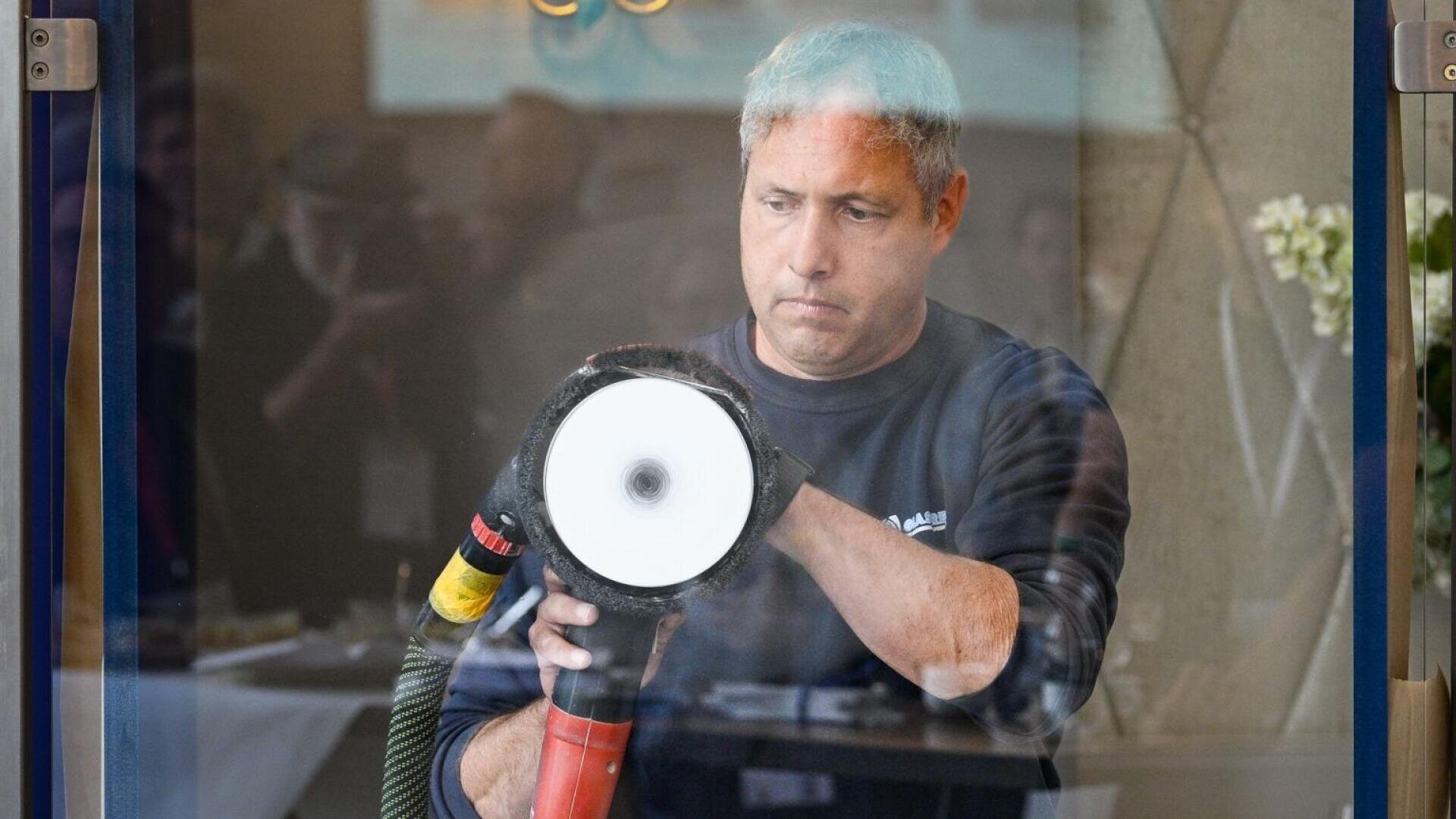 Ein Mann mit ergrauendem Haar poliert mit einer großen Poliermaschine eine Glasoberfläche und konzentriert sich dabei auf seine Arbeit. Das Foto ist von der anderen Seite des Glases aufgenommen und zeigt Spiegelungen und Innendekor.