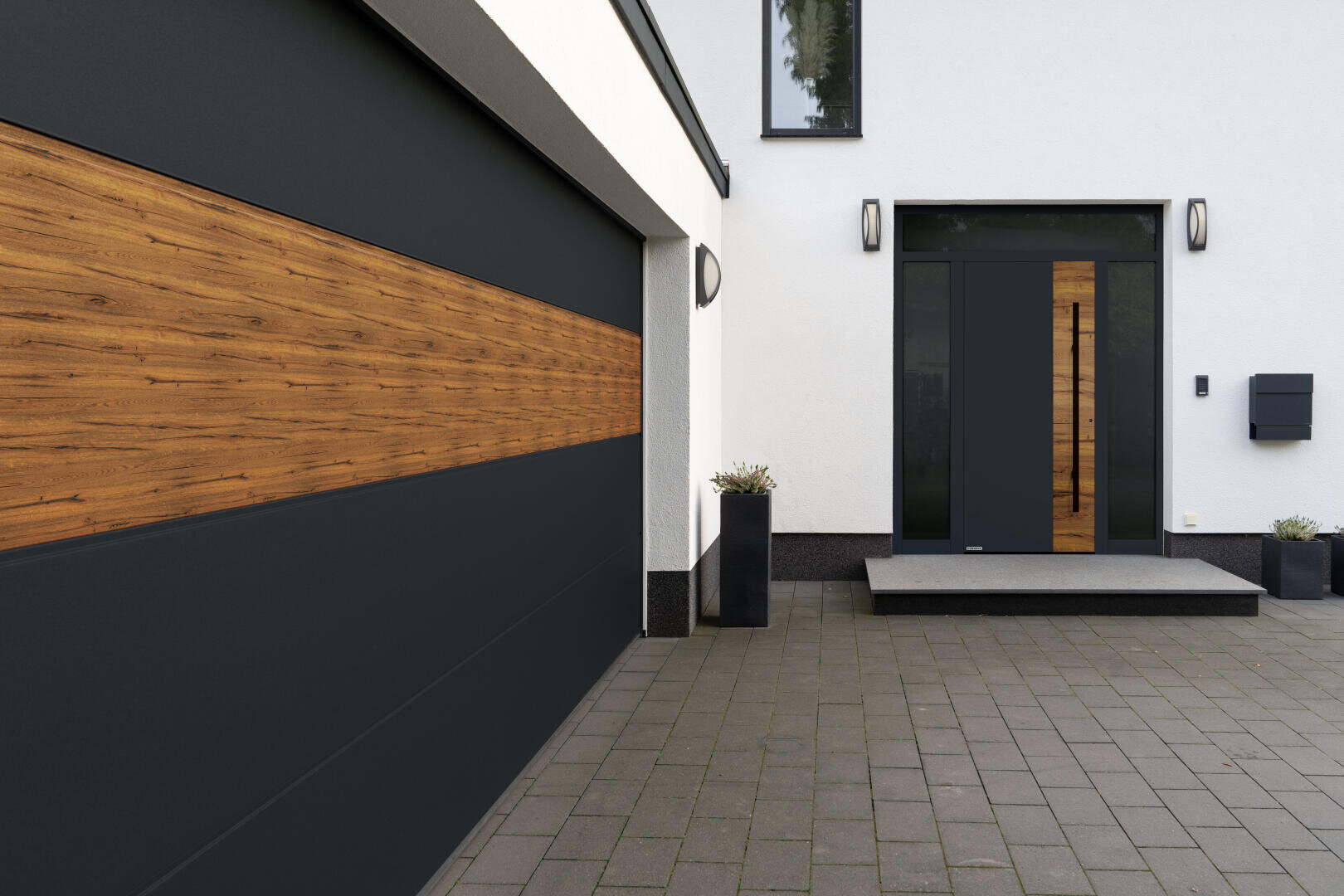 Moderne Hausfassade mit grauem Garagentor und Haustür, beide mit horizontalen Holzpaneelen. Der Eingang ist mit Topfpflanzen, zwei Wandleuchten und einer erhöhten Betonstufe ausgestattet. Die Wände sind weiß mit schwarzer Umrandung.