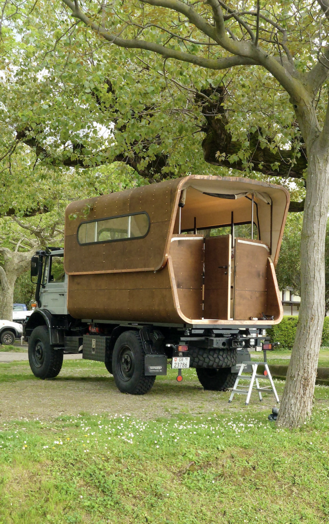 Ein Lastwagen mit einem einzigartigen hölzernen Wohnmobil auf der Ladefläche ist unter Bäumen geparkt. Die Hecktür des Wohnwagens ist offen und gibt den Blick auf das Innere frei, und eine kleine Leiter ermöglicht den Zugang.