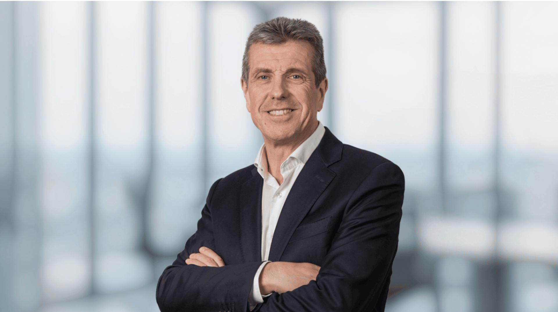 Un homme d'âge moyen vêtu d'un costume sombre et d'une chemise blanche se tient debout, les bras croisés, souriant, devant un arrière-plan de bureau flou avec de grandes fenêtres.