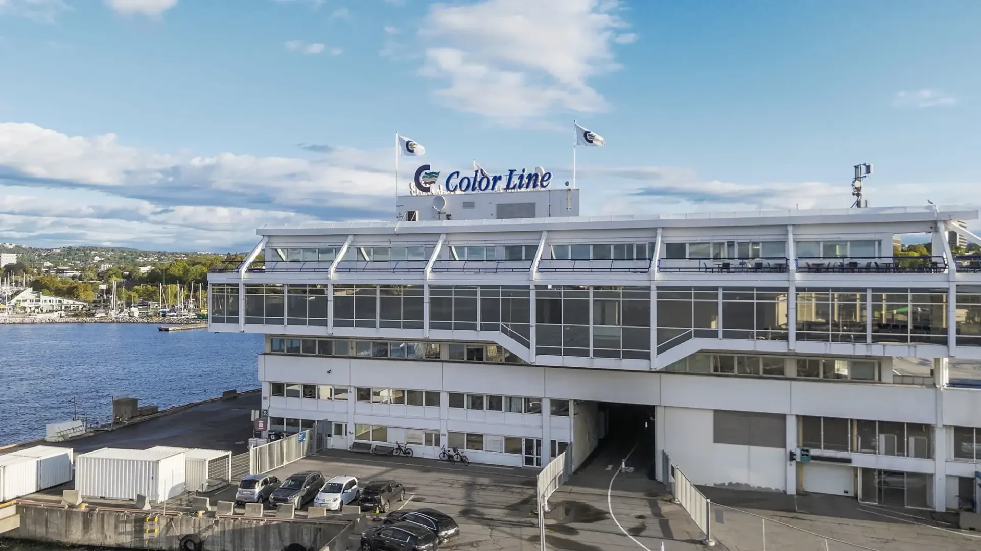 Ein großes, modernes Fährterminalgebäude mit "Color Line"-Schriftzug auf dem Dach, direkt am Wasser gelegen, mit geparkten Autos davor und Blick auf das Wasser und entfernte Grünflächen unter einem teilweise bewölkten Himmel.