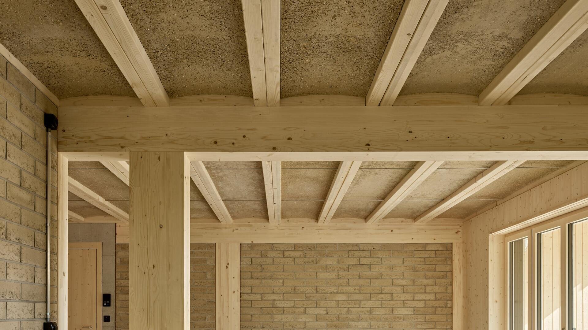 Innenraum eines modernen Zimmers mit freiliegenden Holzbalken, hellem Holzrahmen und beigen Ziegelwänden, wobei durch große Fenster auf der rechten Seite natürliches Licht einfällt.