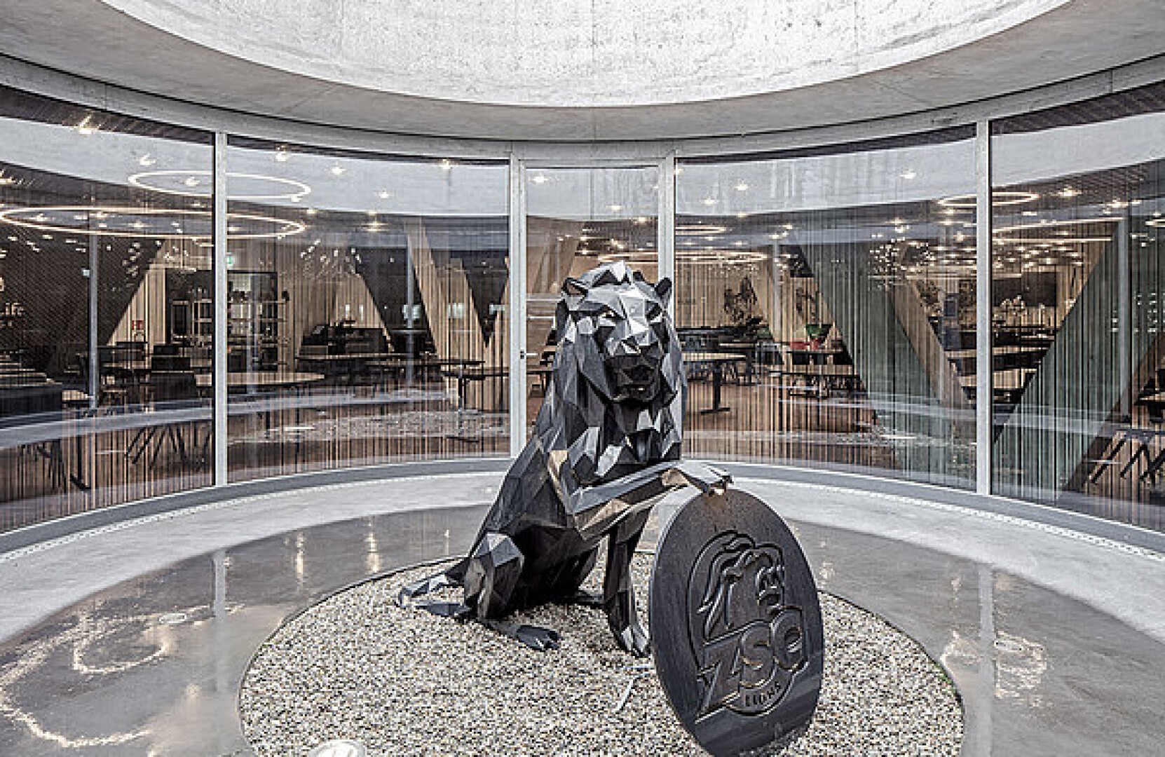 Eine geometrische schwarze Löwenskulptur steht auf Kieselsteinen in einem modernen, runden Raum mit Glaswänden und hält eine runde Plakette mit einem Löwenemblem und den Buchstaben ZSO. Im Hintergrund ist ein stilvolles Interieur mit Tischen und Stühlen zu sehen.