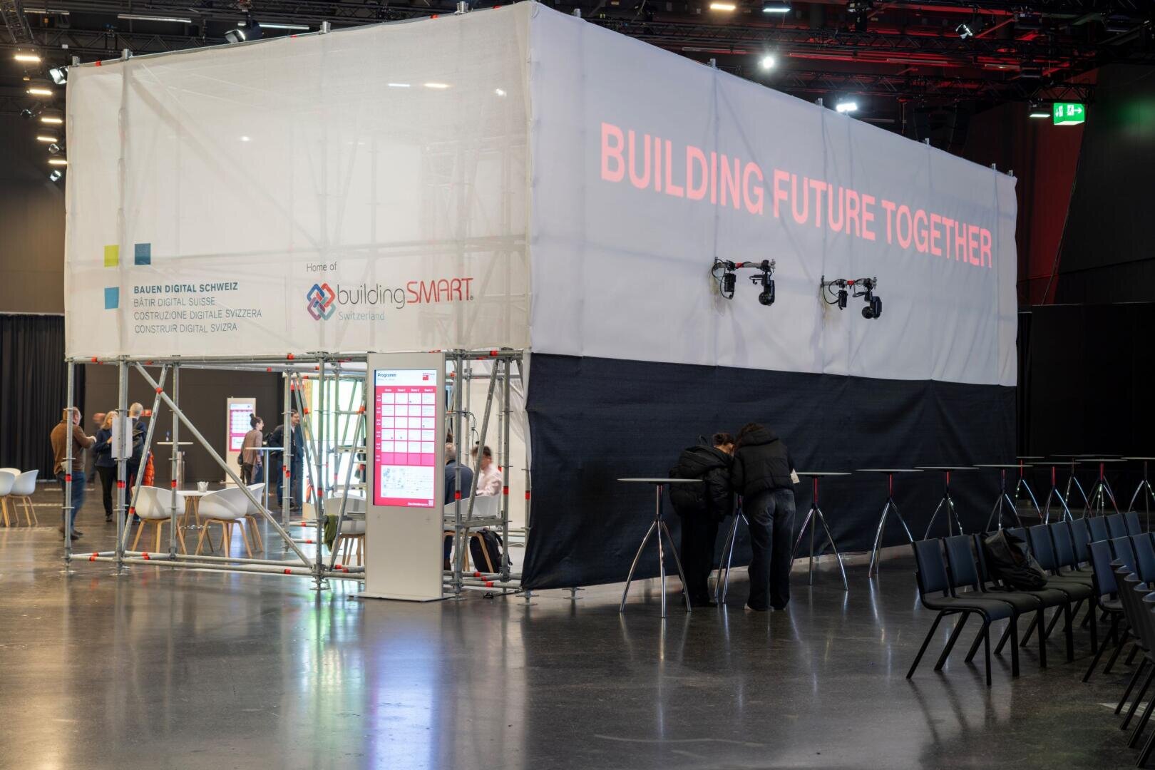 Ein großer weißer Stand mit der Aufschrift BUILDING FUTURE TOGETHER in Rot steht auf einer Veranstaltung in einer Halle. In der Nähe des Standes interagieren Menschen, und auf der rechten Seite sind leere Stühle aufgestellt.