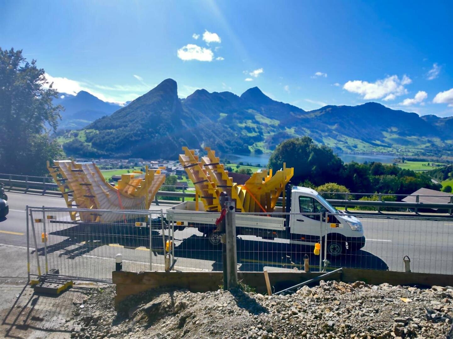 Ein weißer Pritschenwagen transportiert große gelbe Baumaterialien entlang einer Bergstraße, mit grünen Hügeln, Bäumen und dramatischen Gipfeln im Hintergrund unter einem strahlend blauen Himmel.
