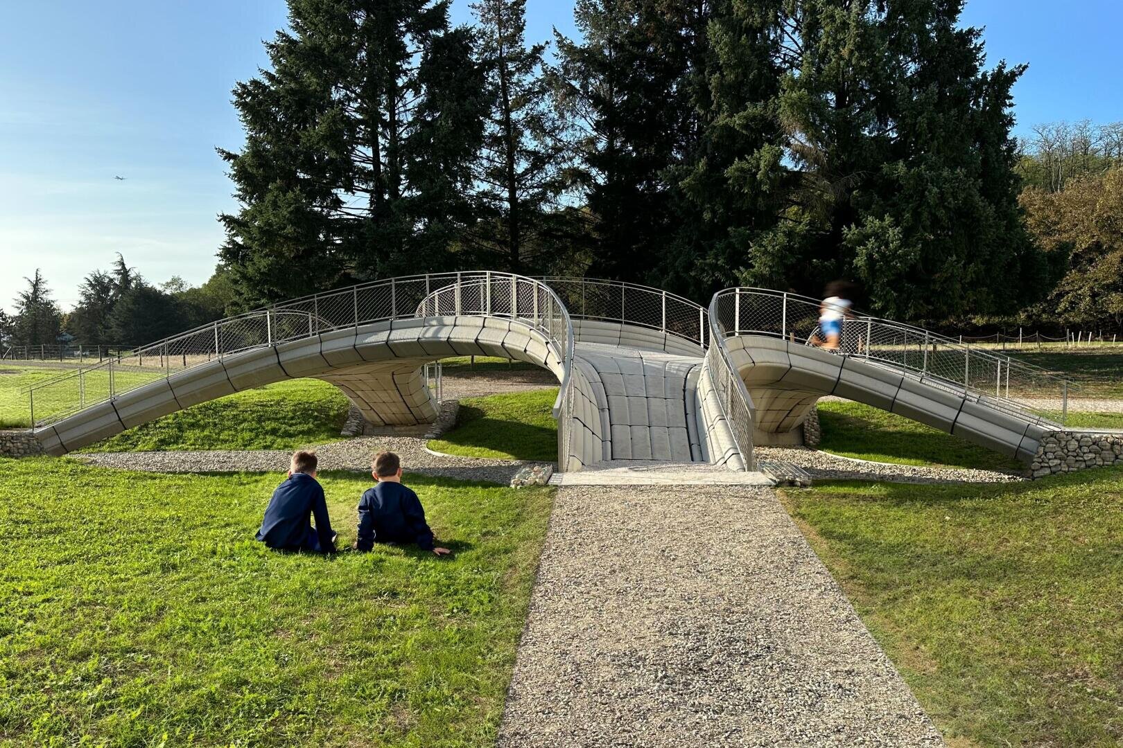 Zwei Kinder sitzen im Gras neben einer einzigartigen, geschwungenen Betonbrücke mit Geländer, über die eine Person in Bewegung läuft. Im Hintergrund sind hohe immergrüne Bäume und ein blauer Himmel zu sehen.