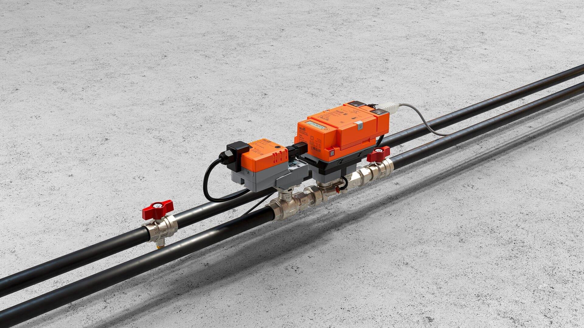 Un gros plan sur des tubes industriels noirs avec des poignées de vannes rouges et des unités de contrôle électroniques orange montées dessus, le tout posé sur un sol en béton gris.