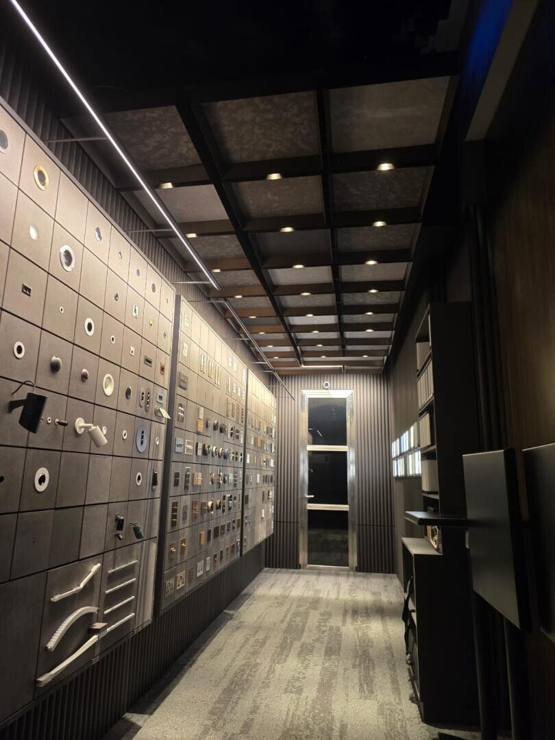 Une salle d'exposition moderne et bien éclairée présente une multitude de ferrures telles que des poignées, des boutons et des robinets, montés de manière ordonnée sur des panneaux muraux gris, avec une moquette au sol et un grand miroir au fond.