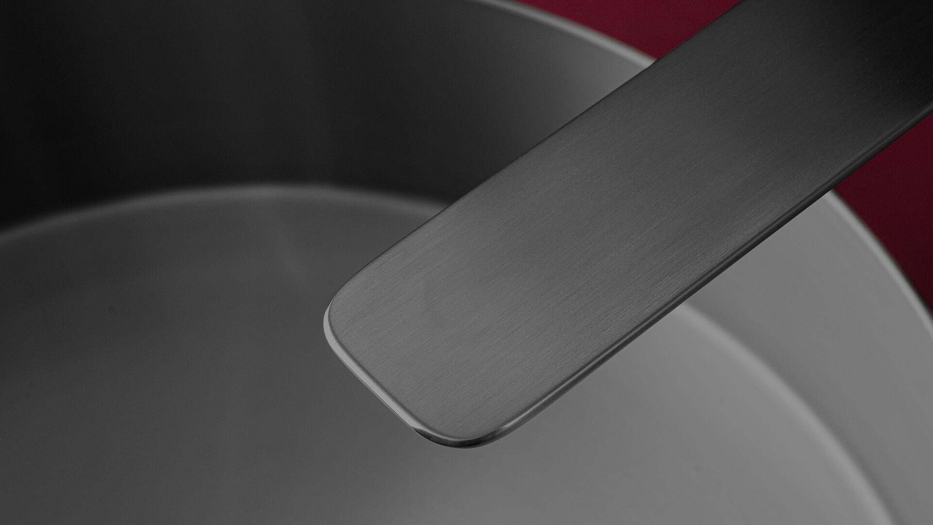 Gros plan d'un pot en acier inoxydable avec une poignée métallique assortie sur un fond rouge profond. L'intérieur du pot et la poignée sont en métal brossé.