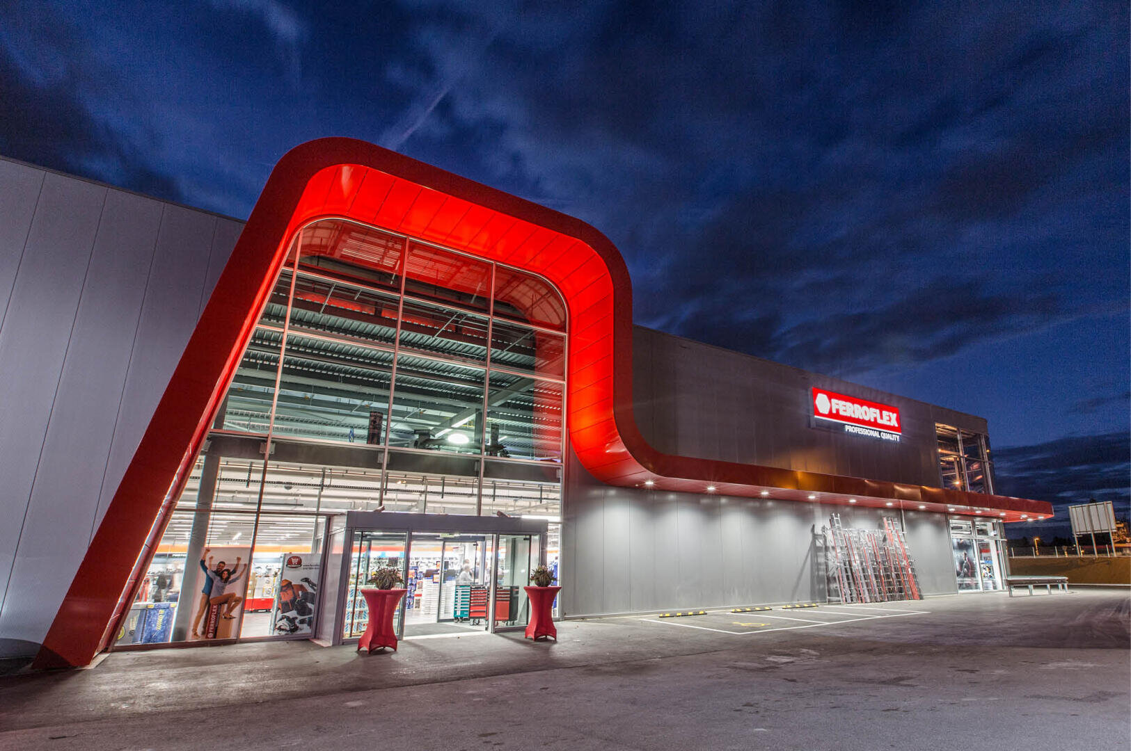 Ein großes, modernes Geschäftsgebäude mit einem auffälligen roten architektonischen Merkmal und einem gläsernen Eingang wird in der Abenddämmerung beleuchtet. Auf dem Schild über dem Eingang steht FERROFLEX. Der Himmel ist dunkel und bewölkt.