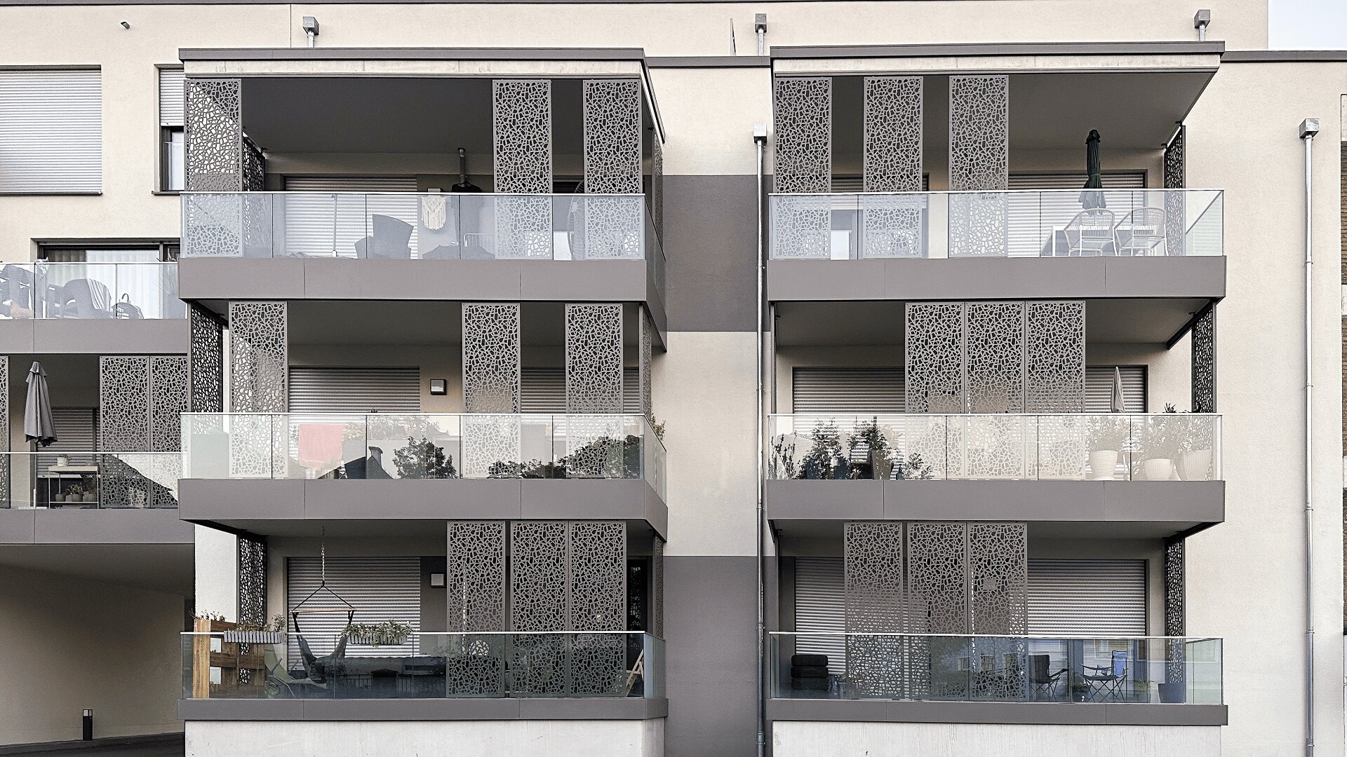 Fassade eines modernen Wohnhauses mit sechs Balkonen, die mit Glasgeländern und dekorativen Metallpaneelen ausgestattet sind. Einige Balkone sind mit Pflanzen, Outdoor-Möbeln und anderen persönlichen Gegenständen geschmückt.