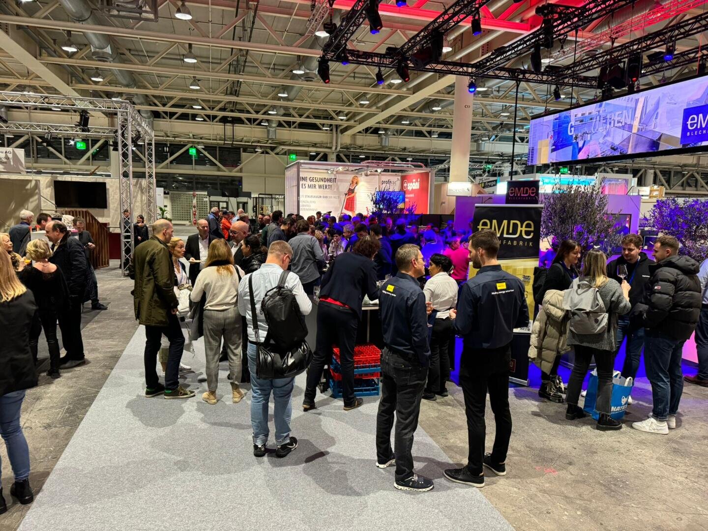 Eine große Gruppe von Menschen versammelt sich auf einer überdachten Ausstellung oder Messe und interagiert in der Nähe verschiedener Stände und Displays bei heller Beleuchtung. Einige Teilnehmer tragen Markenjacken, und im Hintergrund sind Banner und Bildschirme zu sehen.