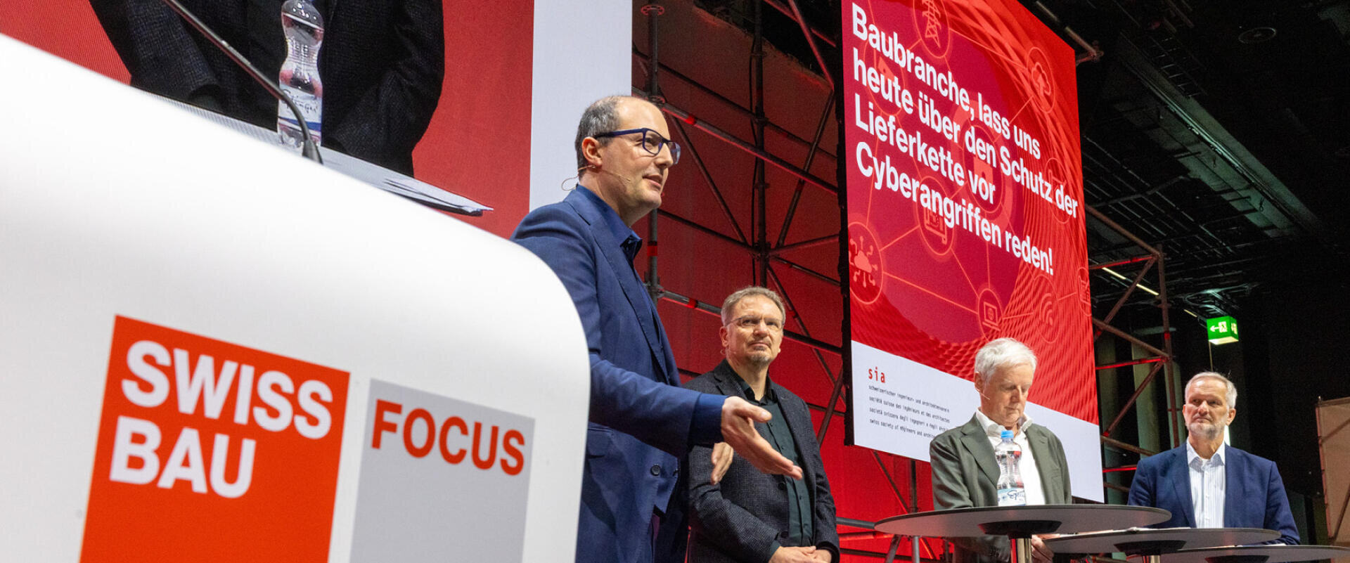 Vier Männer stehen auf einer Bühne an einer Swissbau Focus Veranstaltung. Ein Mann spricht, während die anderen zuhören. Im Hintergrund zeigt ein roter Bildschirm Texte über Bauwesen, Lieferketten und Cyber-Risiken an. Im Vordergrund steht ein Swissbau Focus-Podium.