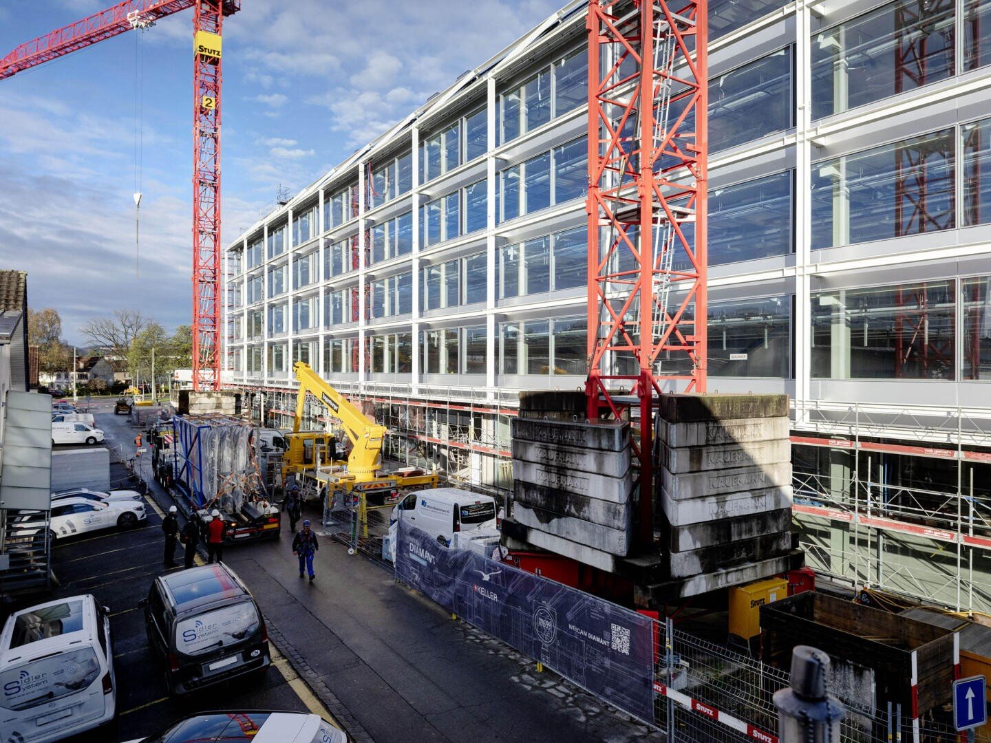 Un chantier avec une grande grue rouge et un bâtiment moderne en verre en construction. Des ouvriers, des véhicules et des équipements sont visibles à côté du bâtiment, sous un ciel partiellement nuageux.