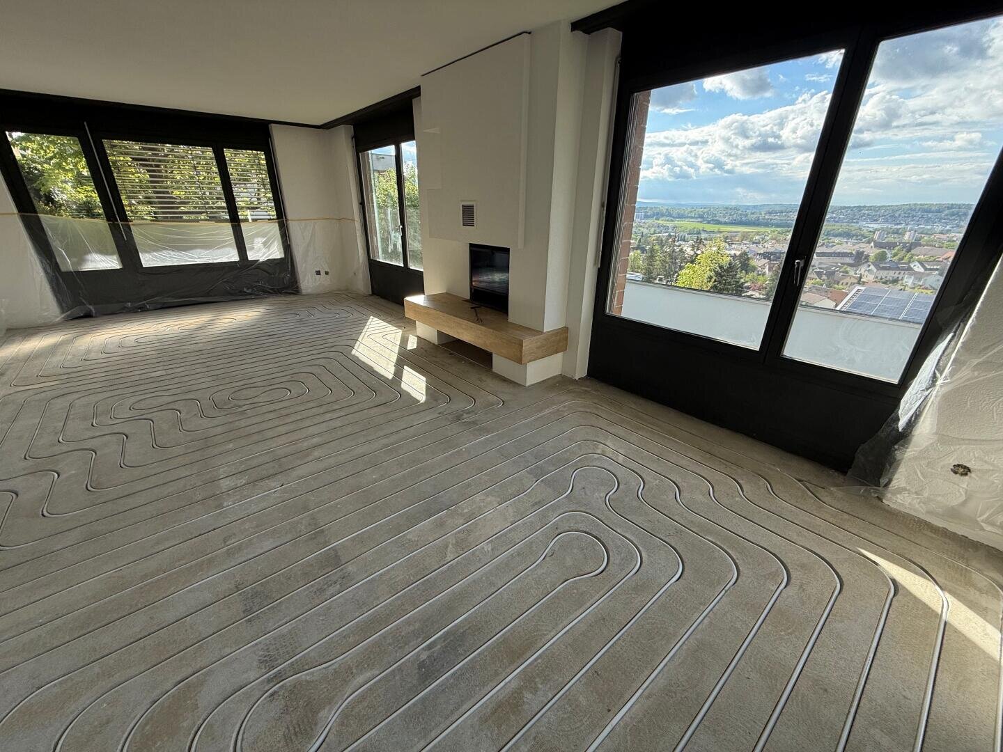 Ein heller Raum mit großen Fenstern und Blick auf die Stadt, mit freiliegenden Fußbodenheizungsrohren, die in den Betonboden eingelassen sind, bevor der endgültige Bodenbelag verlegt wird. Teile der Fenster und Wände sind zum Schutz mit Kunststoffplatten abgedeckt.