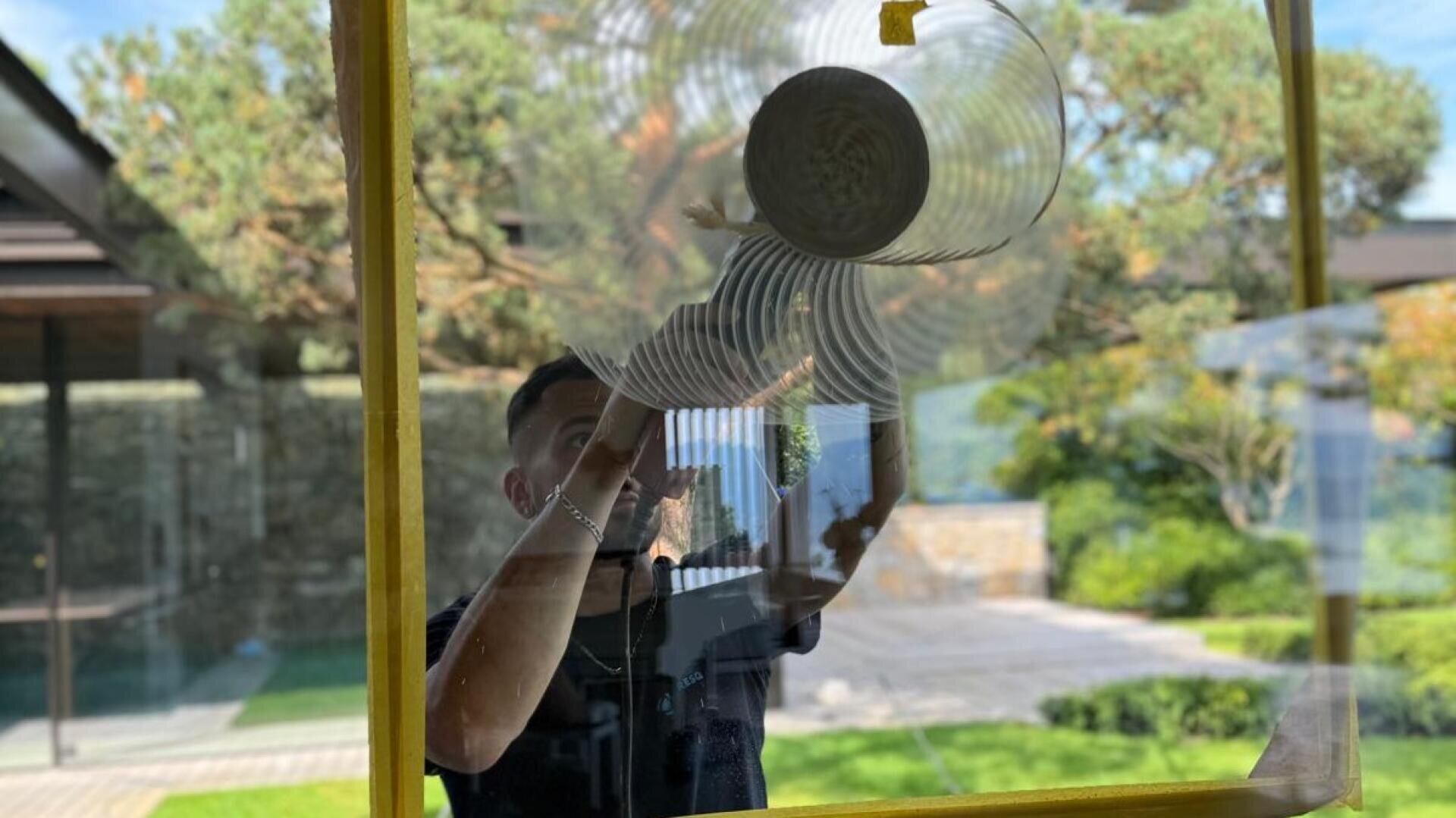 Eine Person reinigt ein großes Fenster mit einer kreisförmigen Bewegung, wodurch wirbelnde Muster auf dem Glas entstehen. Durch das Fenster sind draußen Grünflächen und ein Hof zu sehen.